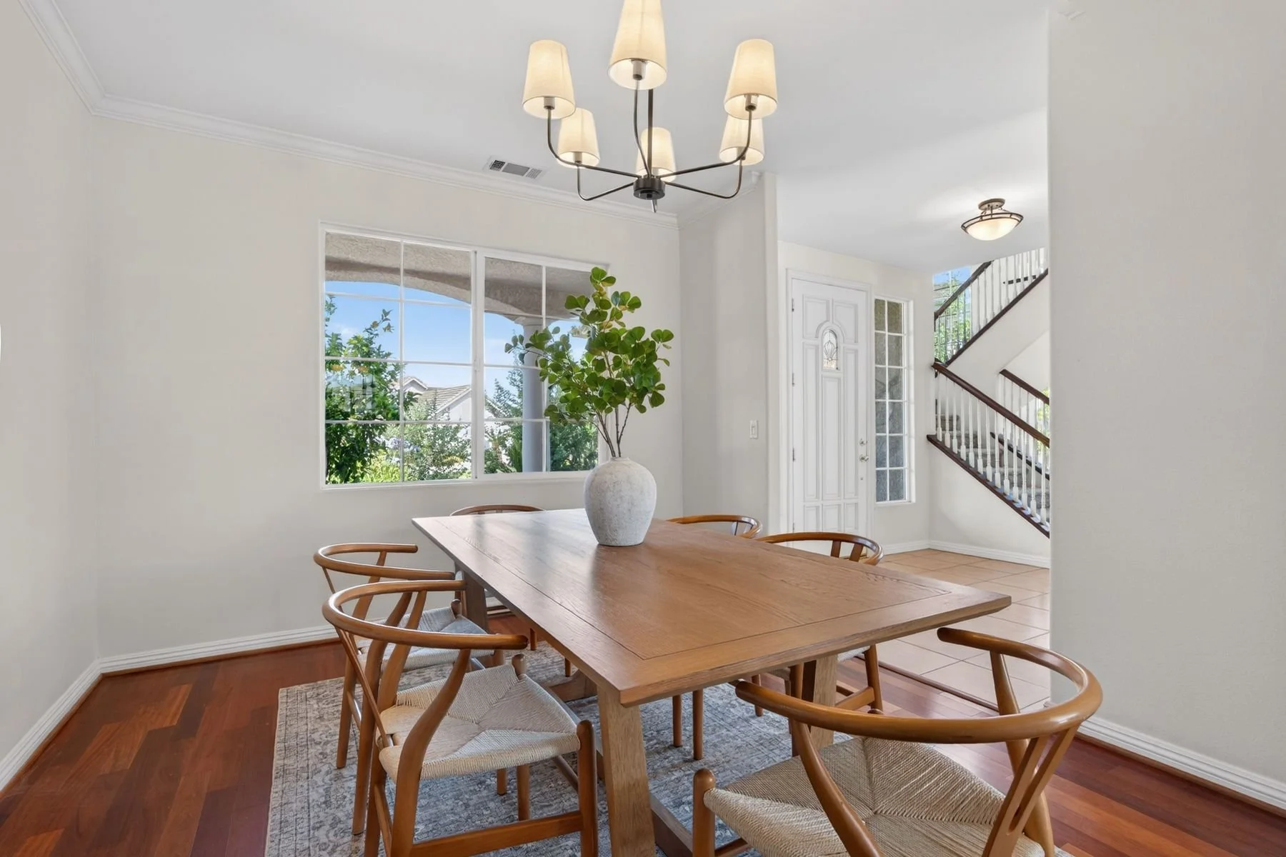 A modern dining room with a wooden table, six wooden chairs with woven seats, a white vase with green foliage, large window showing greenery outside, staircase with wooden handrail, entry door with glass panels, and a chandelier with eight lampshades