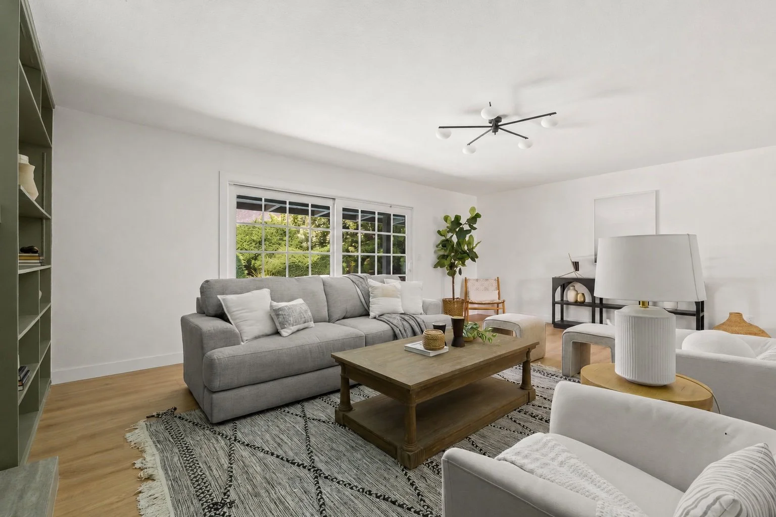 Living room with white walls, large window, gray sofa with white and gray pillows, wooden coffee table, white armchair, lamp, potted plant, and decor accents.