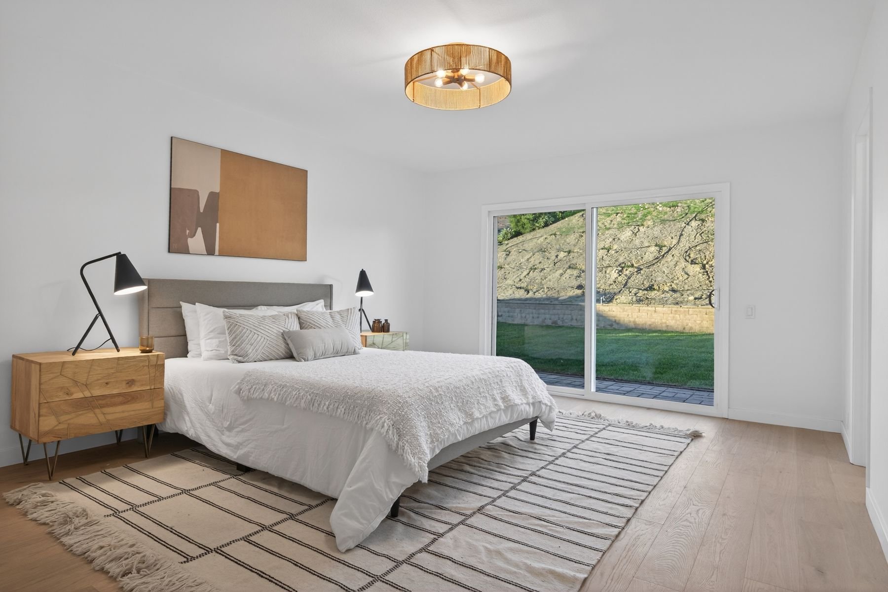 A modern bedroom with a large bed, white bedding, and multiple pillows, flanked by black cone-shaped lamps on wooden nightstands, a striped rug on light hardwood floors, a large window showing a grassy outdoor area and a stone wall, and a ceiling lig
