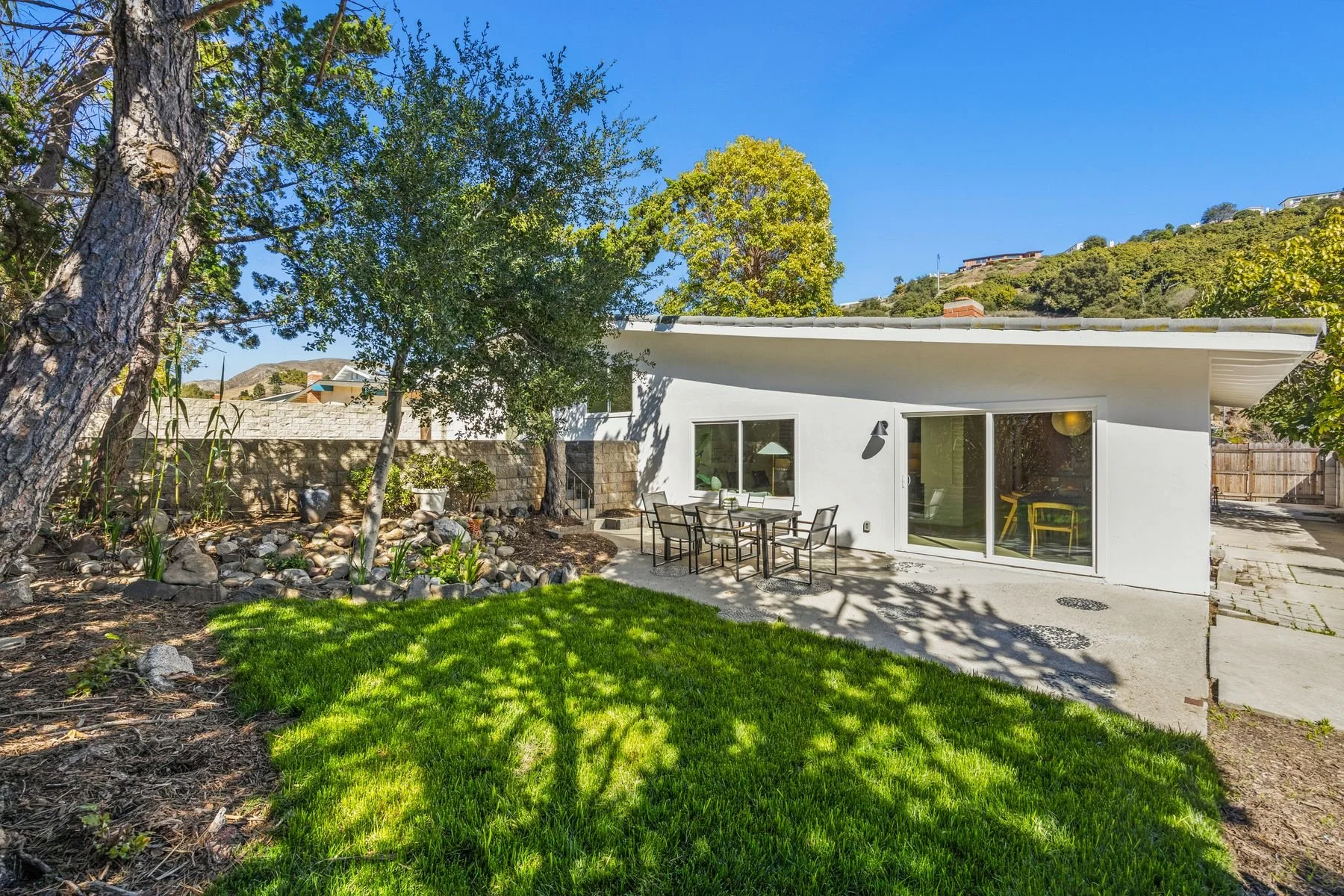 A modern white house with large sliding glass doors, an outdoor dining table with six chairs, surrounded by a landscaped backyard with rocks, small plants, and a grassy area, under a clear blue sky with trees and hills in the background.