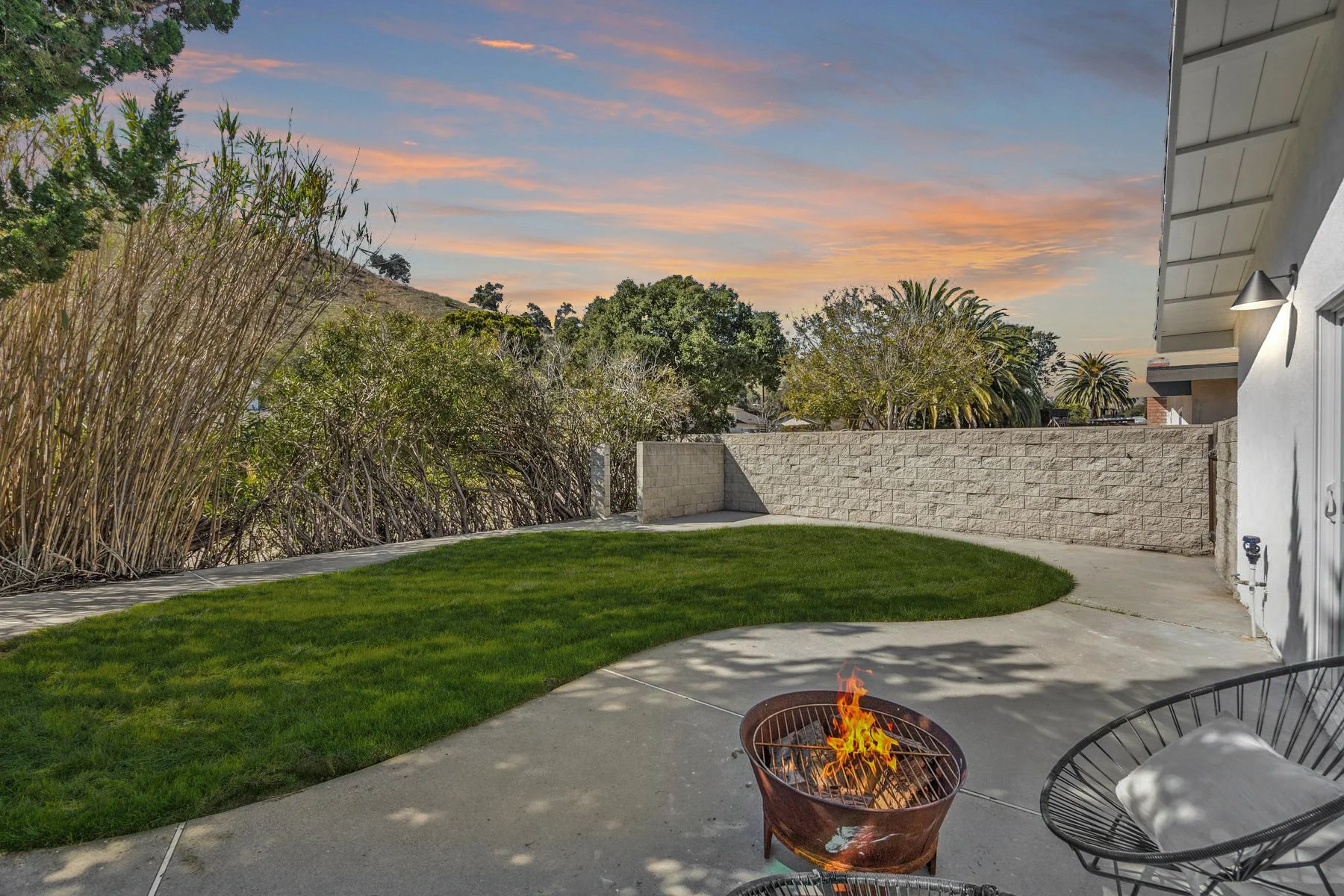 A backyard at sunset with a grassy lawn, concrete patio, a fire pit with flames, outdoor chairs, a white house wall, and a stone fence. There are trees and bushes in the background, with a colorful sunset sky.