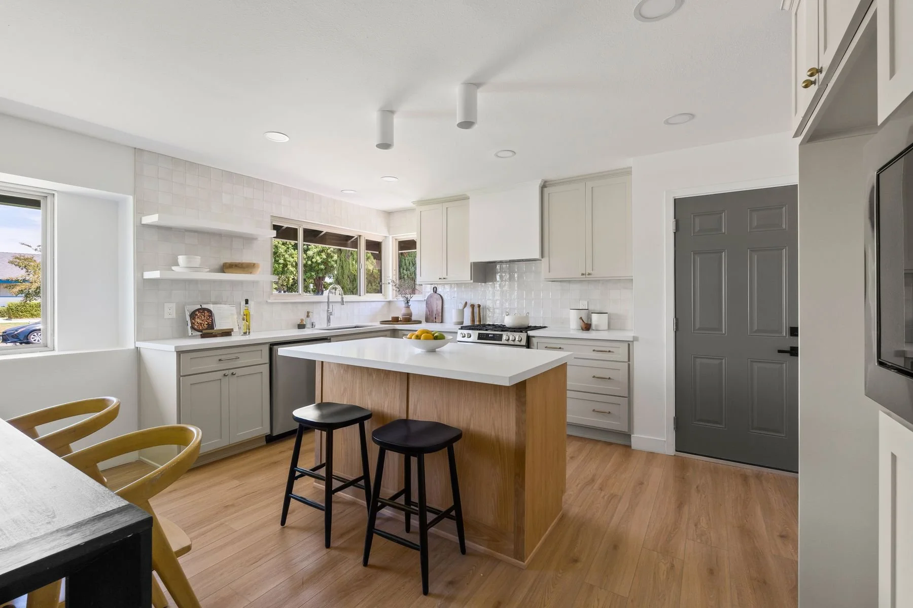 Modern bright kitchen with white cabinets, a white countertop island, and wood flooring. There are large windows showing trees outside. The kitchen has stainless steel appliances, open shelves with bowls, and a bowl of fruit on the island. Bar stools