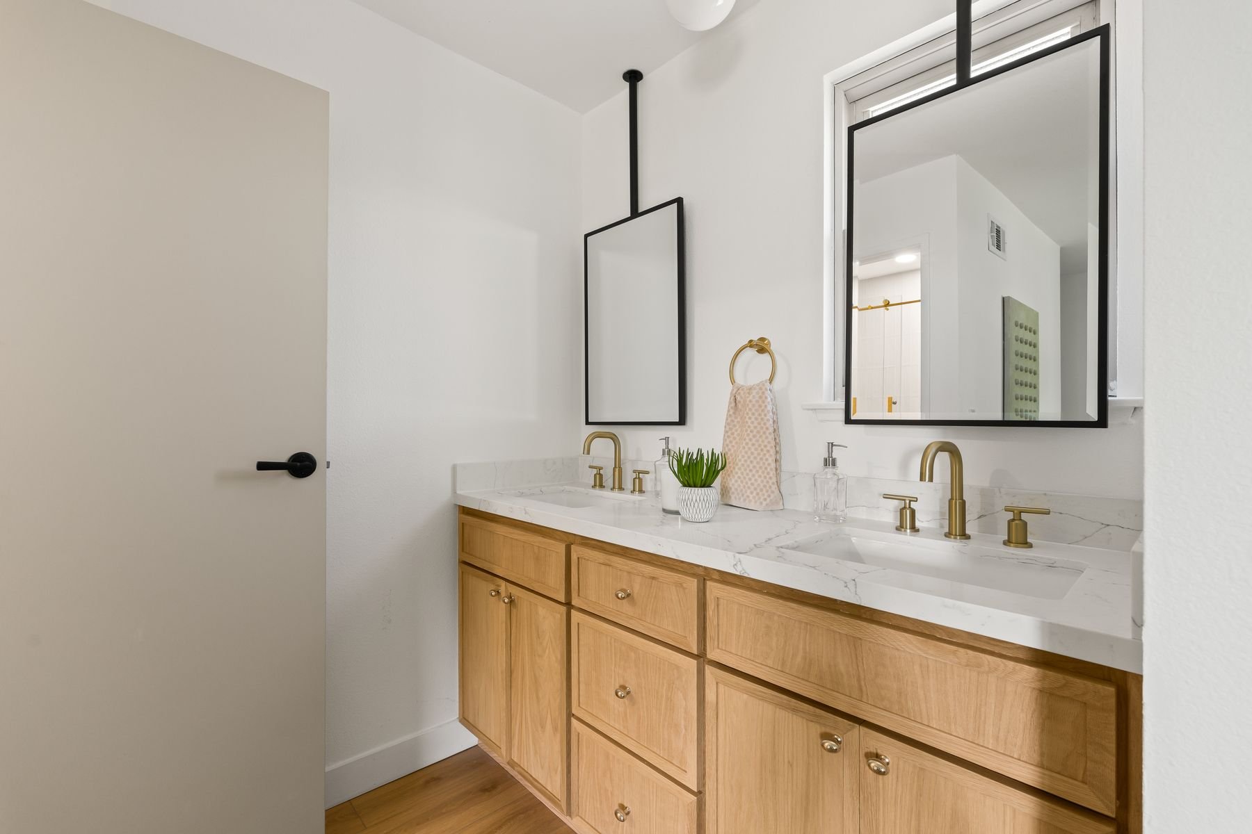 Bathroom vanity with light wood cabinets, marble countertop, two gold faucets, mirror, window with natural light, decorative items including a plant and towel, white walls.