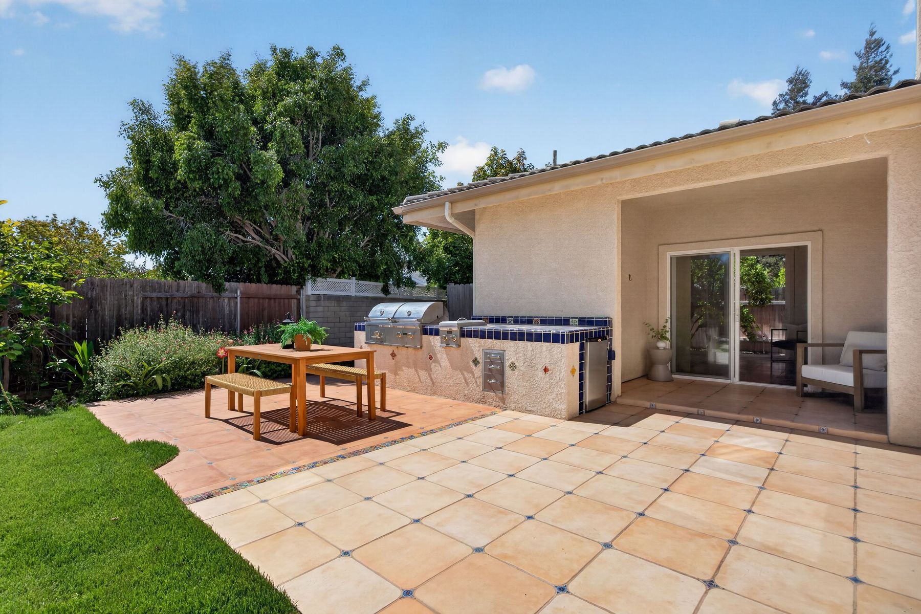 Backyard patio with a wooden table and two benches, a built-in barbecue, a sliding glass door leading to the house, a sitting chair, potted plants, lush green trees, and a wooden fence, under a clear blue sky.