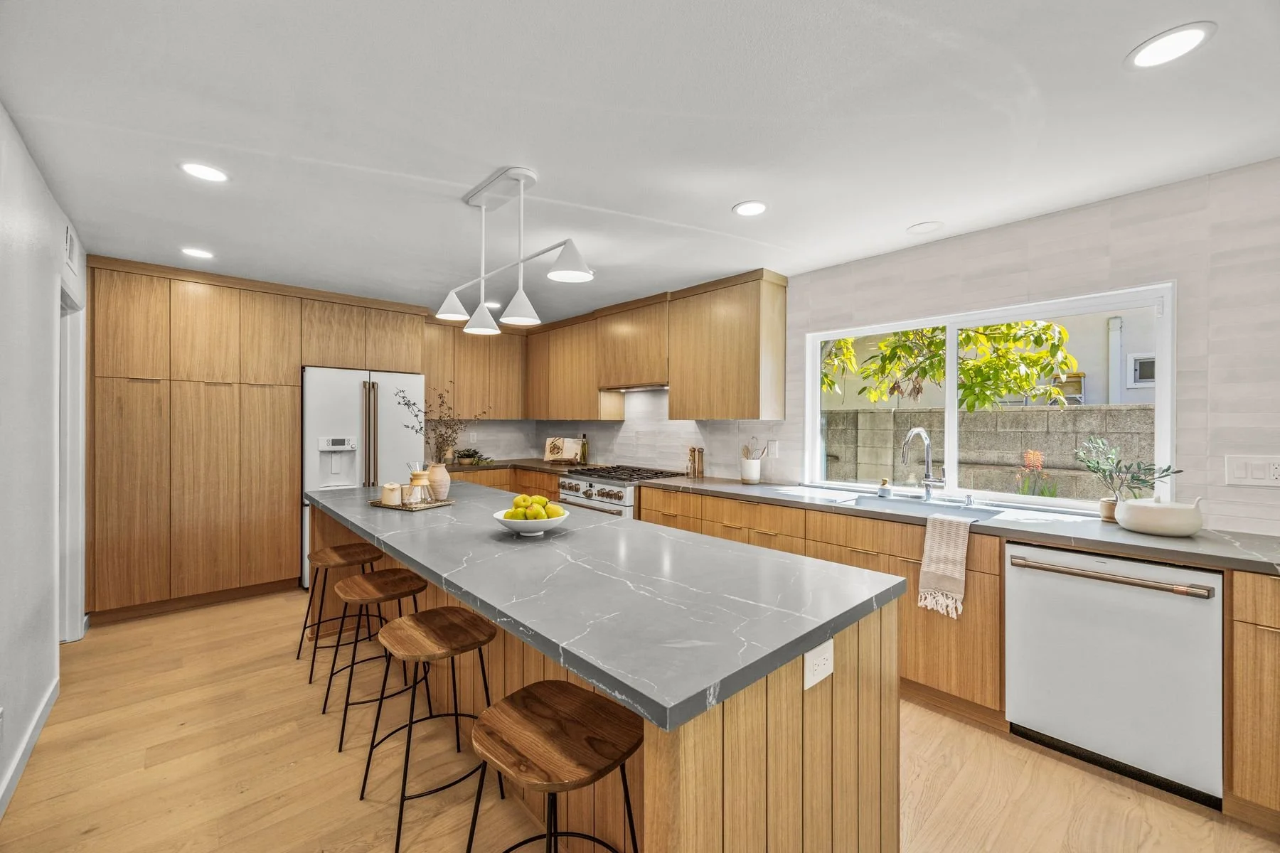 Modern kitchen with wooden cabinets, a marble-topped island, a large window showing outdoor greenery, and pendant lighting.