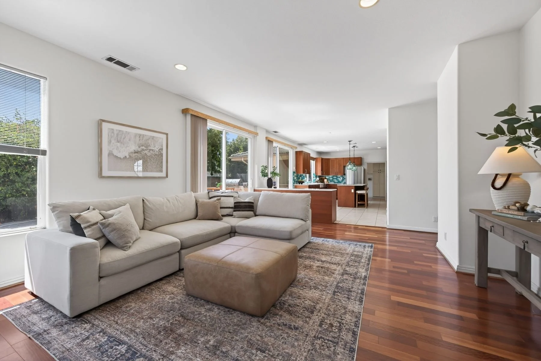 Living room with white couch, pillows, and ottoman, large windows, and open kitchen in the background with wooden cabinets and a tile backsplash.