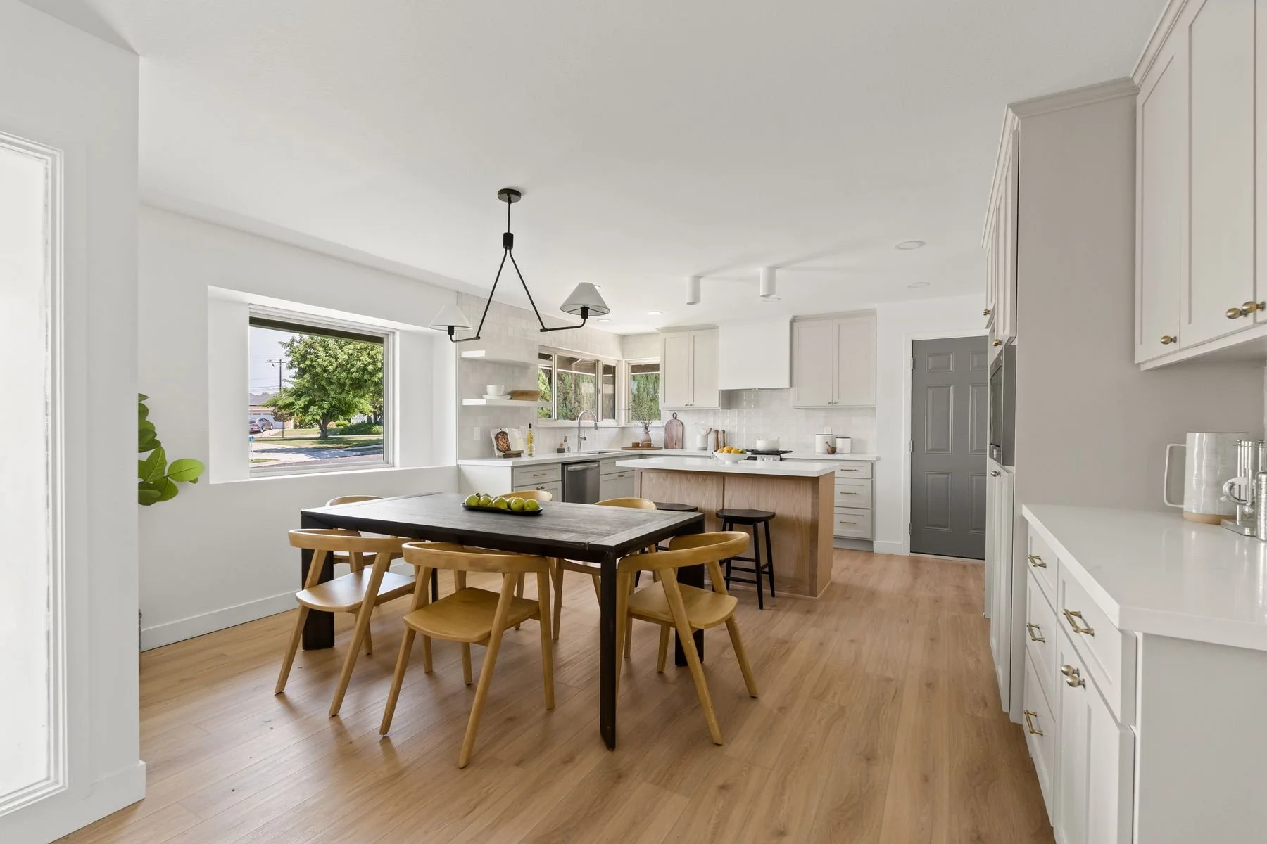 Modern kitchen with white cabinetry, gray door, wooden dining table with six chairs, hanging light fixture, window showing a tree and street view, and various kitchen items like bowls, fruit, and appliances.
