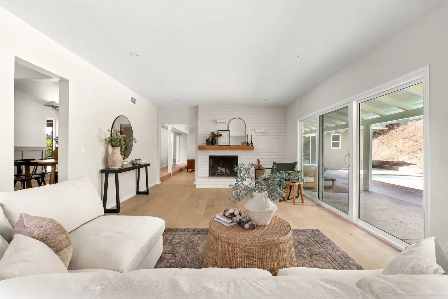Bright living room with white walls, a white sofa, a round wicker coffee table with a large vase and greenery, a white brick fireplace with decor on the mantel, sliding glass doors leading to an outdoor patio, and hardwood floors.