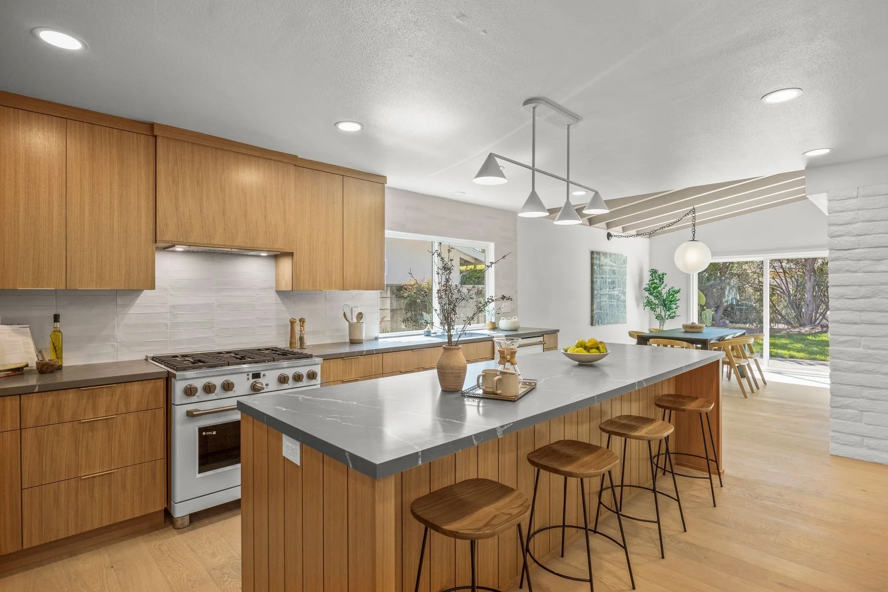 Modern kitchen with wooden cabinets, a white stove, and a large island with a gray marble countertop. There are four wooden barstools at the island. The space has natural light from a window and sliding glass door, with plants and a dining area visib