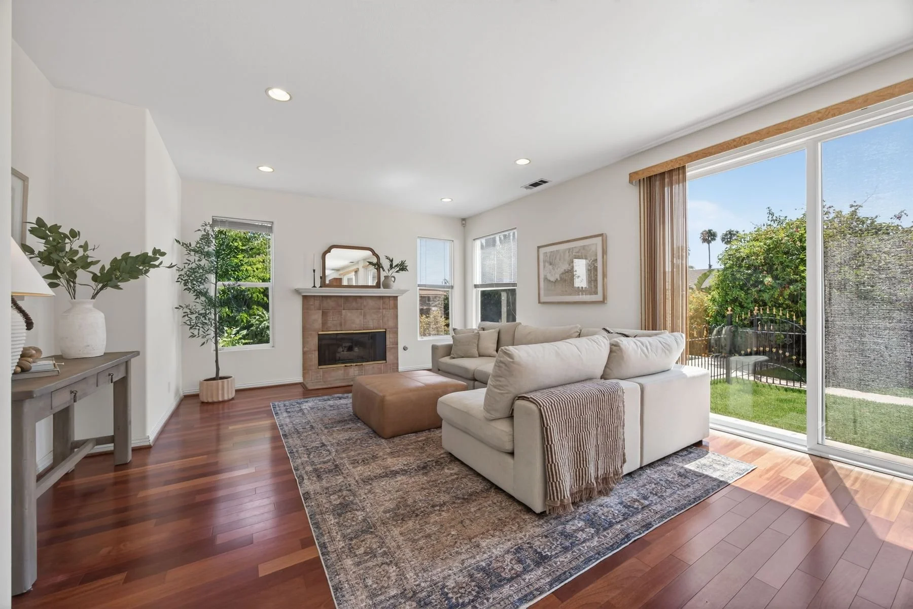 Living room with beige sofas, fireplace, hardwood floors, and large sliding glass doors showing a backyard with trees and a tall fence.