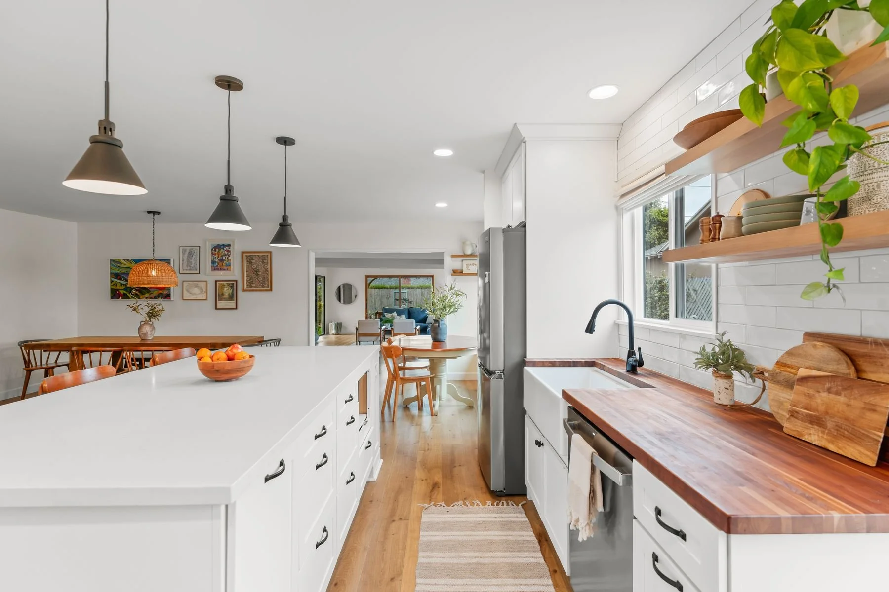 Open-concept kitchen with white cabinets, a large white island, wooden countertops, and open shelving with dishes and plants. Visible dining area with wooden table and artwork on the wall, adjacent living space with seating and windows showing outdoo