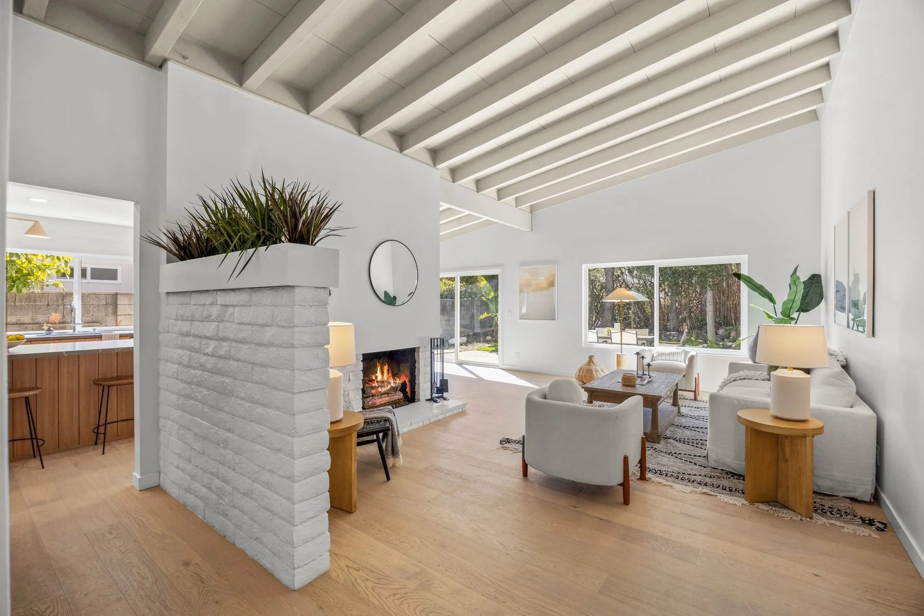 Bright living room with white walls and a vaulted ceiling featuring exposed beams. There is a white brick fireplace with a fire burning, flanked by small tables with lamps. A wooden coffee table with decorative items sits on a patterned rug. Large wi