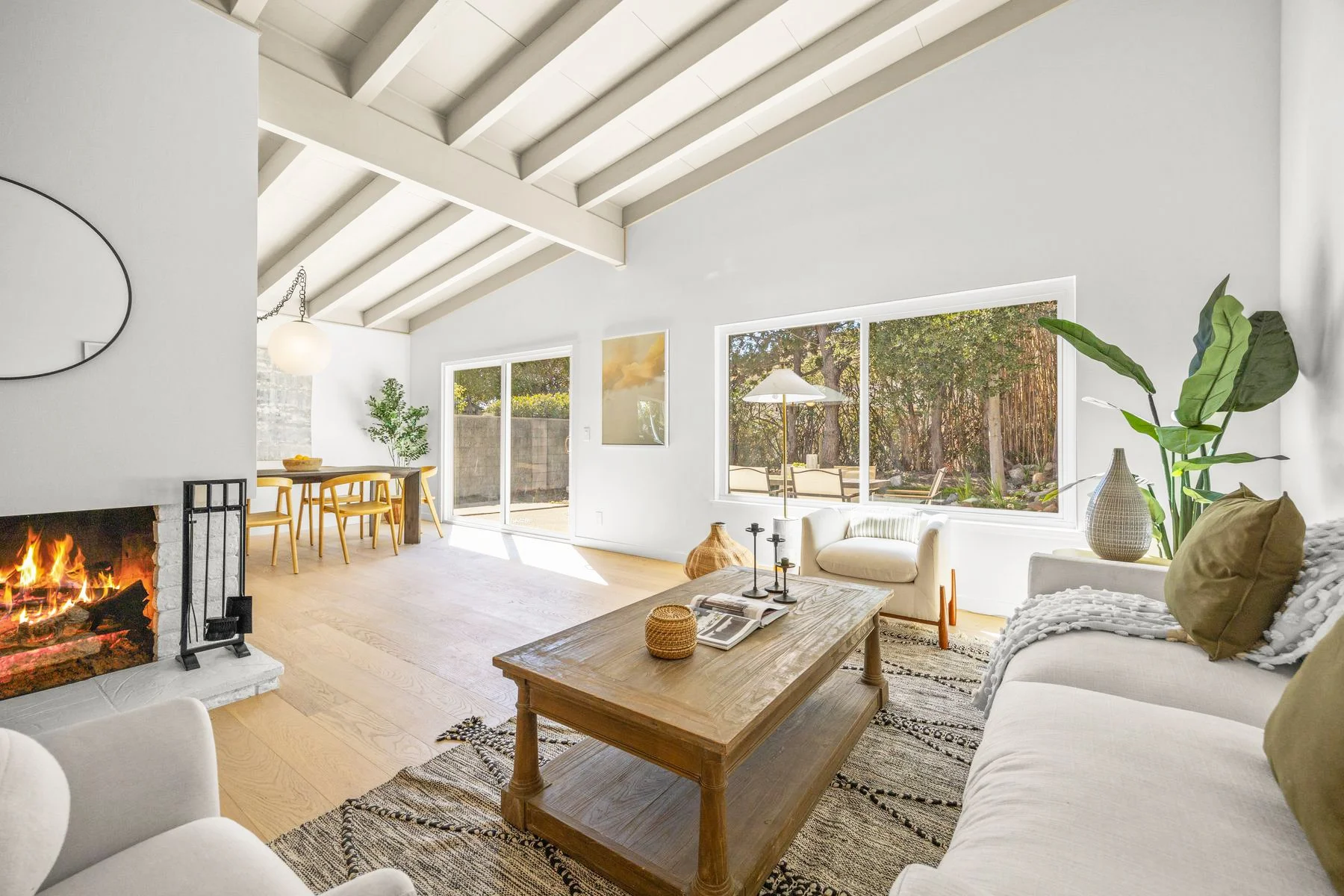 Living room with white walls, large windows, a fireplace, cream sofa, wooden coffee table, and plants.