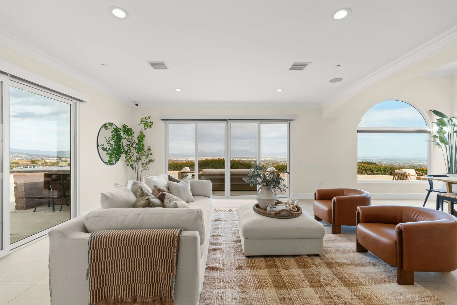 Living room with white sofa, brown leather chairs, sliding glass door and large window showing outdoor view, green plants, and decorative items.