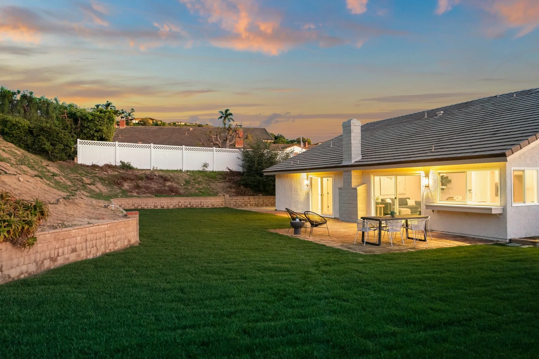 A backyard view during sunset showing a well-maintained green lawn, a patio with outdoor seating, a white house with large windows illuminated from inside, and a white fence with neighboring houses and trees in the background under a colorful sky.