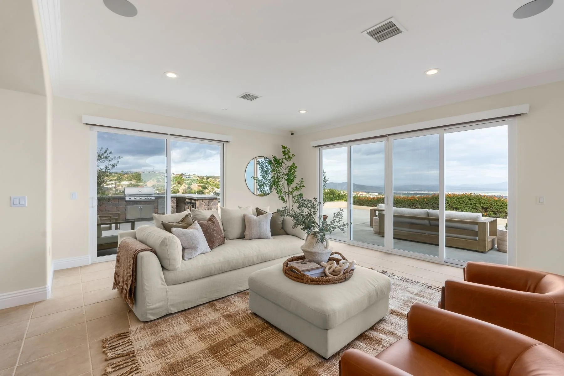 Living room with large windows showing outdoor view, beige sofa with pillows, a round mirror, and potted plant. Patio with lounge chairs and barbecue visible outside.