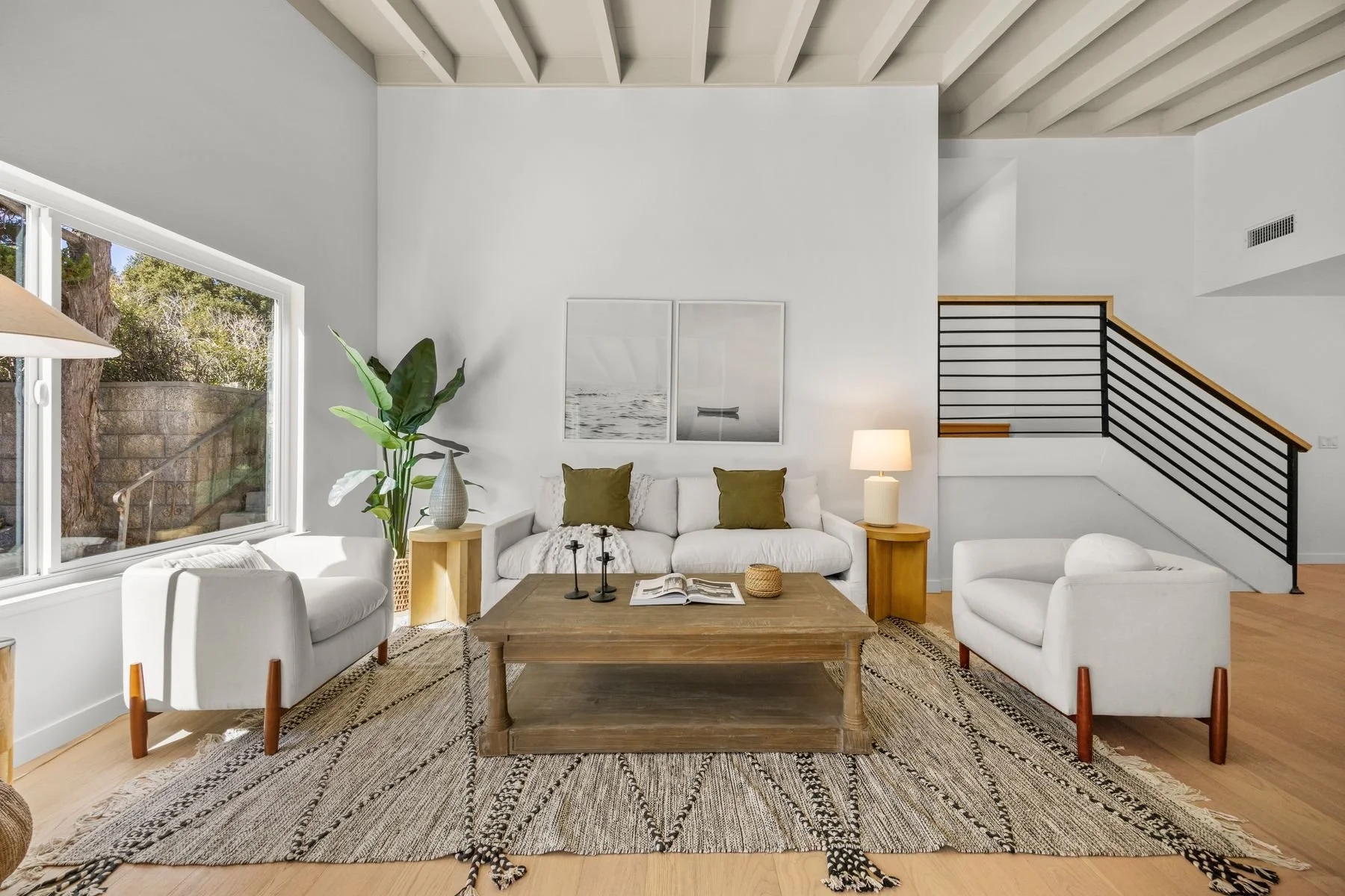 Living room with white couches, a wooden coffee table, and artwork above the sofa. Large window with greenery outside, a tall plant in the corner, and a staircase with a black railing on the right. neutral color scheme, natural light, and decorative 