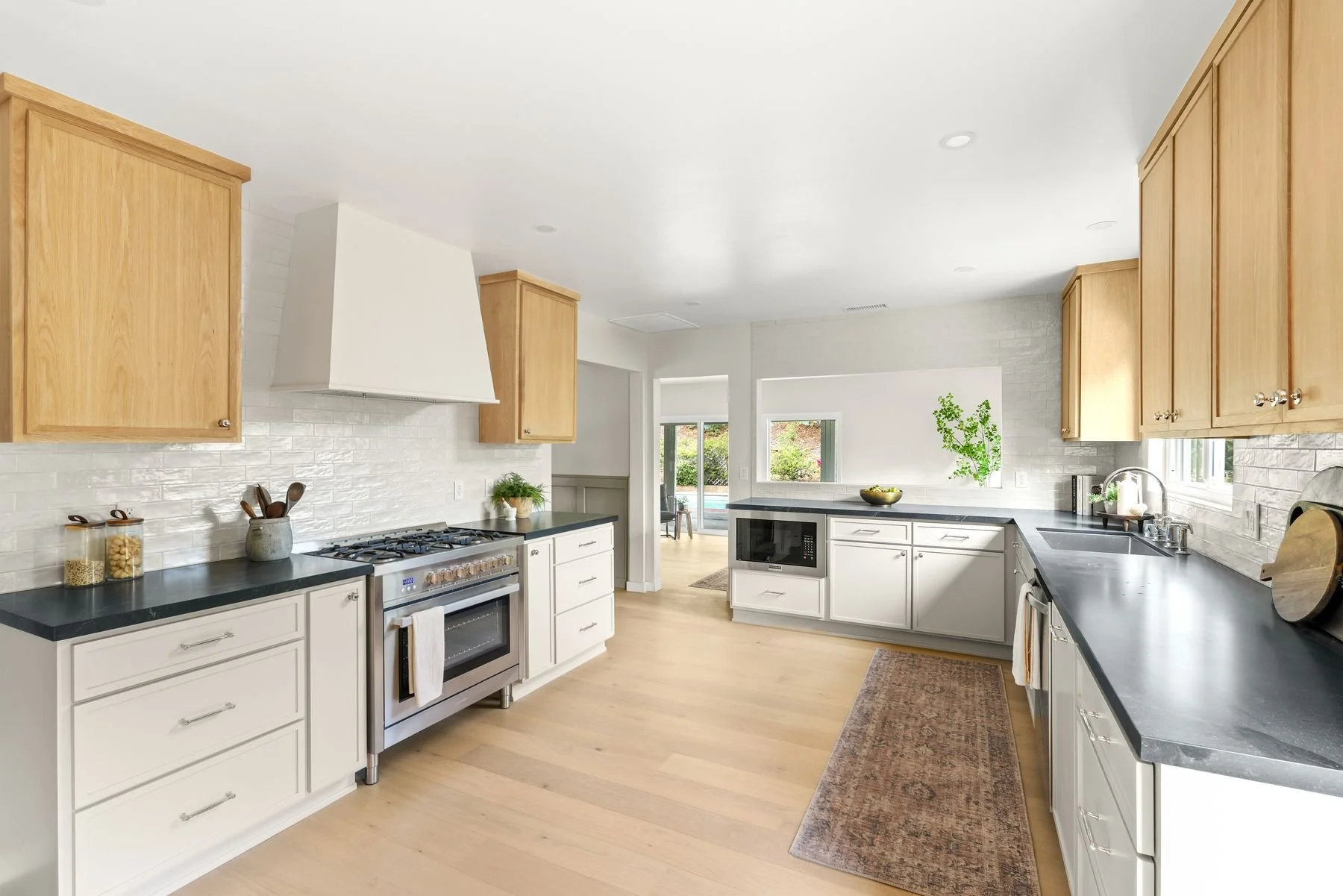Modern kitchen with white cabinets, black countertops, stainless steel stove, and wooden upper cabinets. Light wood flooring and potted plants near windows.
