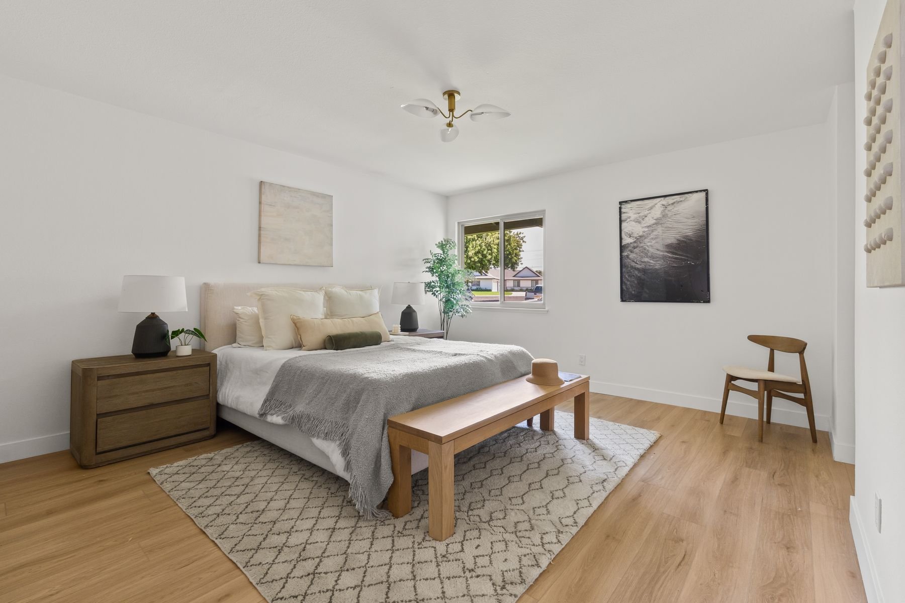 A bedroom with a large bed covered with white and gray bedding, wooden nightstands with lamps and plants, a window with trees outside, black and white artwork on the walls, a wooden bench at the foot of the bed, a chair in the corner, and light wood 
