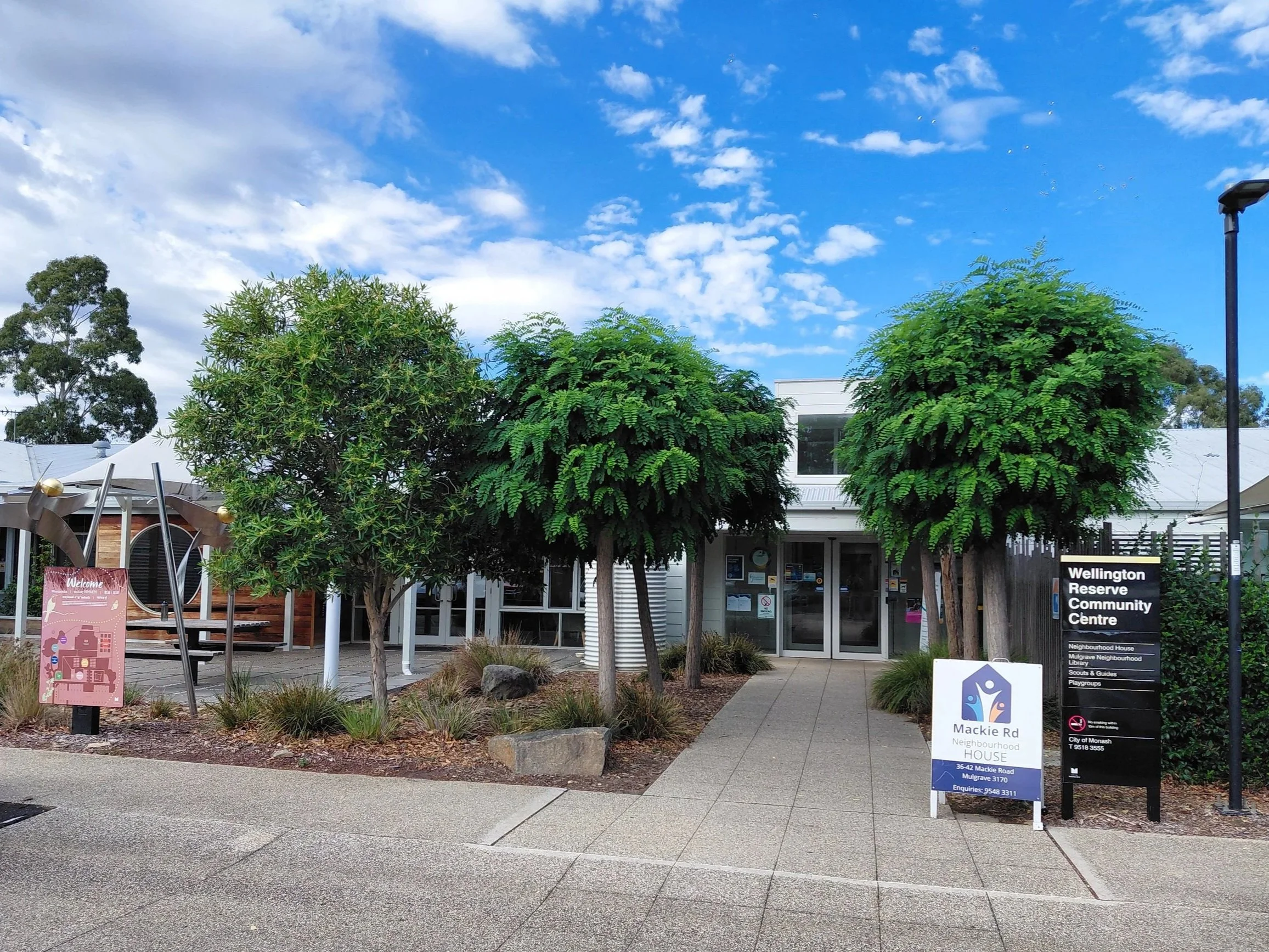 Front street view of Mackie Road Neighbourhood House in Mulgrave of a paved walkway between tree lined gardens leading to the double front doors at the entrance.  Outdoor table setting under shade sail covering, permanent signage and A frame
