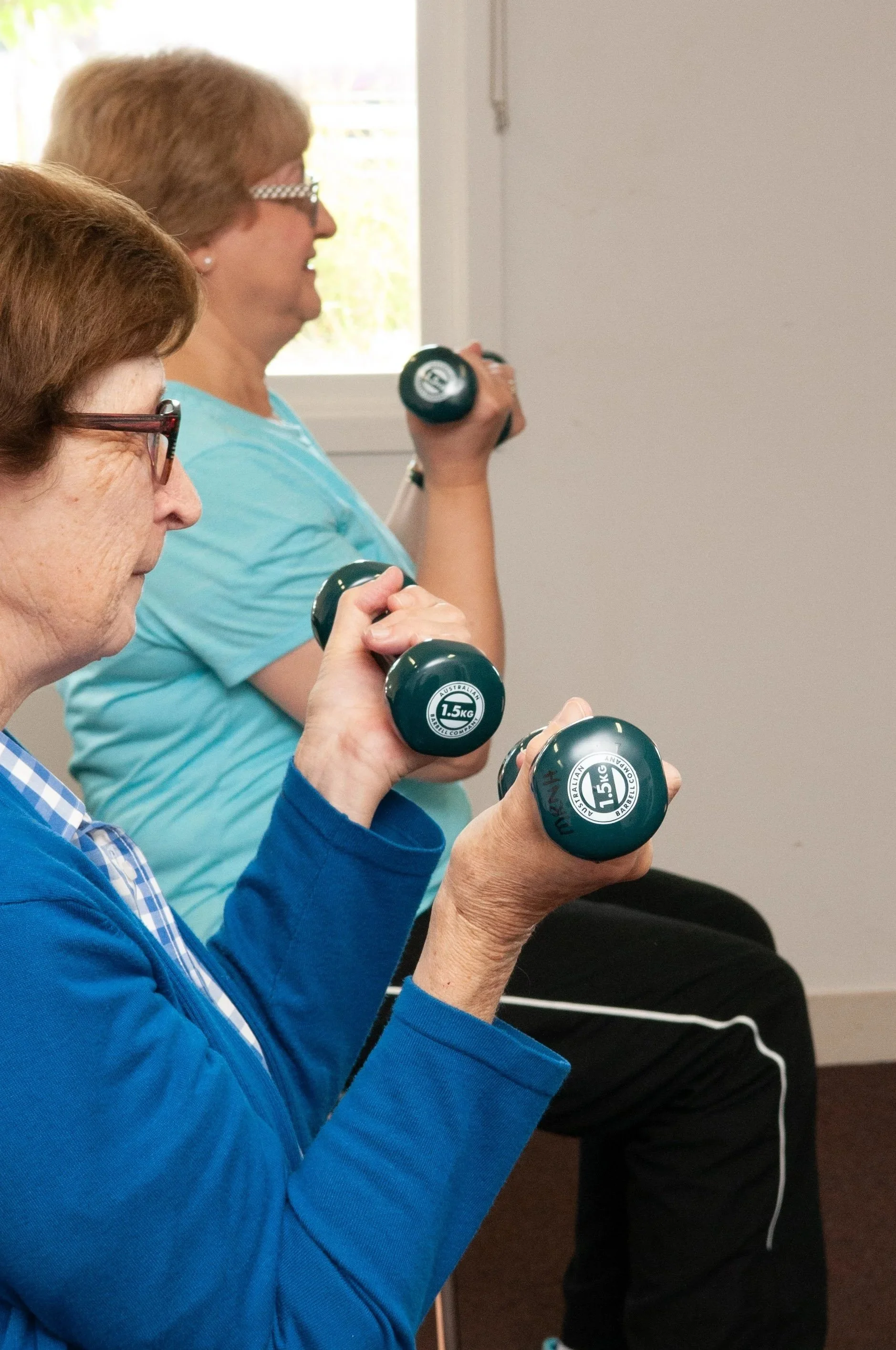 Two older women holding dumbbells and facing towards a fitness instructor off screen