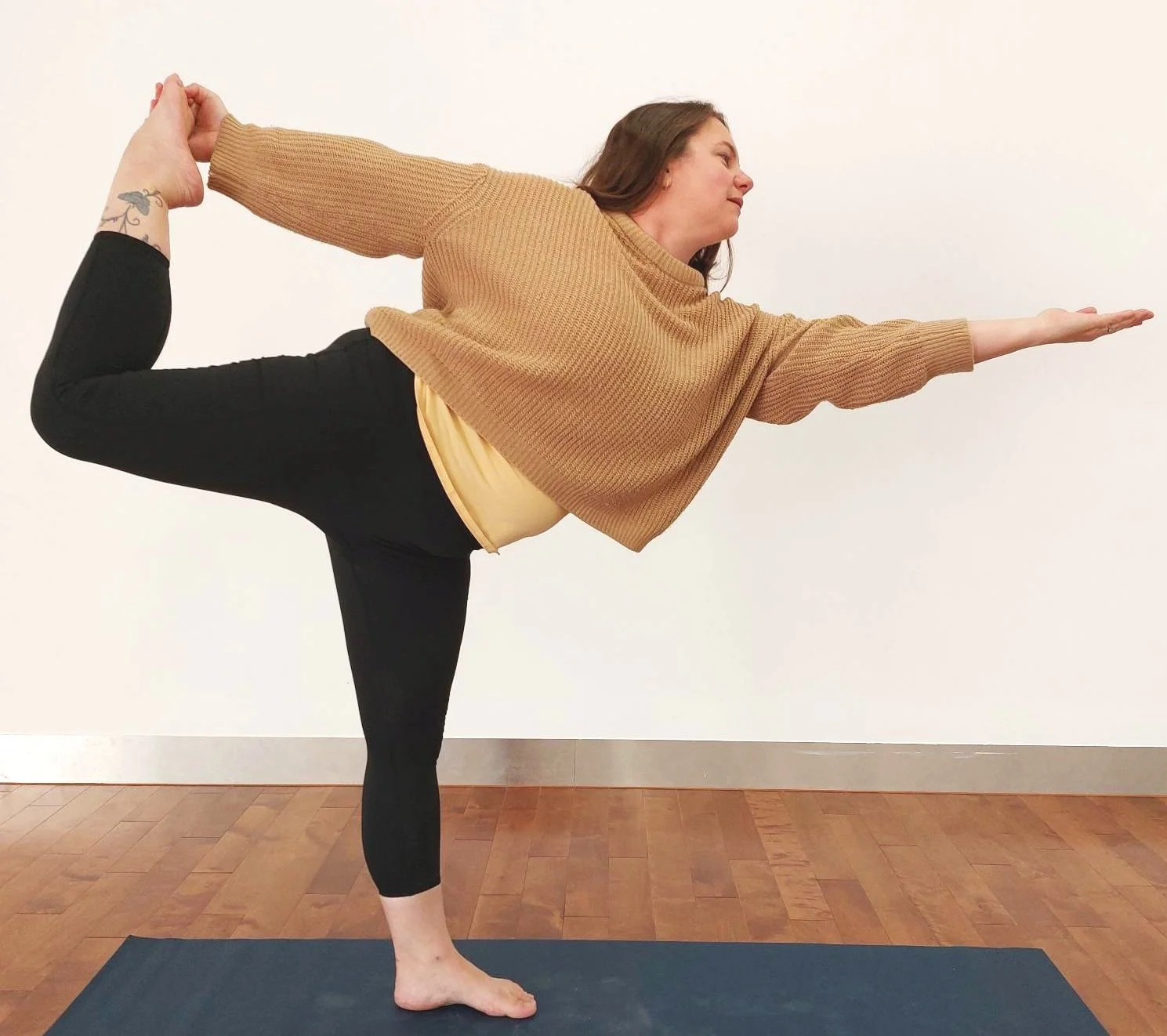 Woman in beige jumper on yoga mat in Dancer Pose standing on left leg while holding right foot behind, leaning forward with left arm outstretched