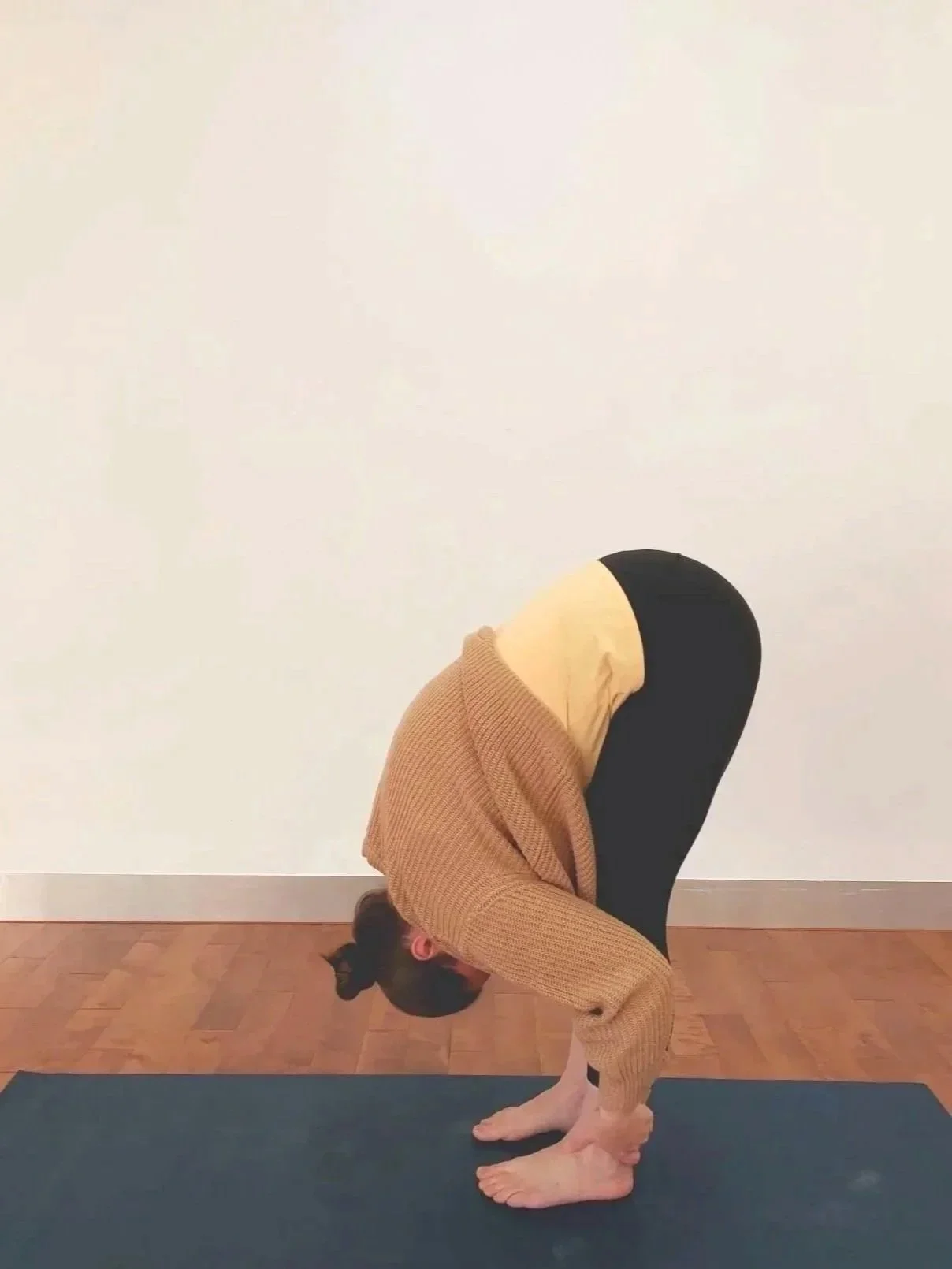 Woman in a beige jumper on a yoga mat in a Forward Fold pose with straight legs, head and torso down