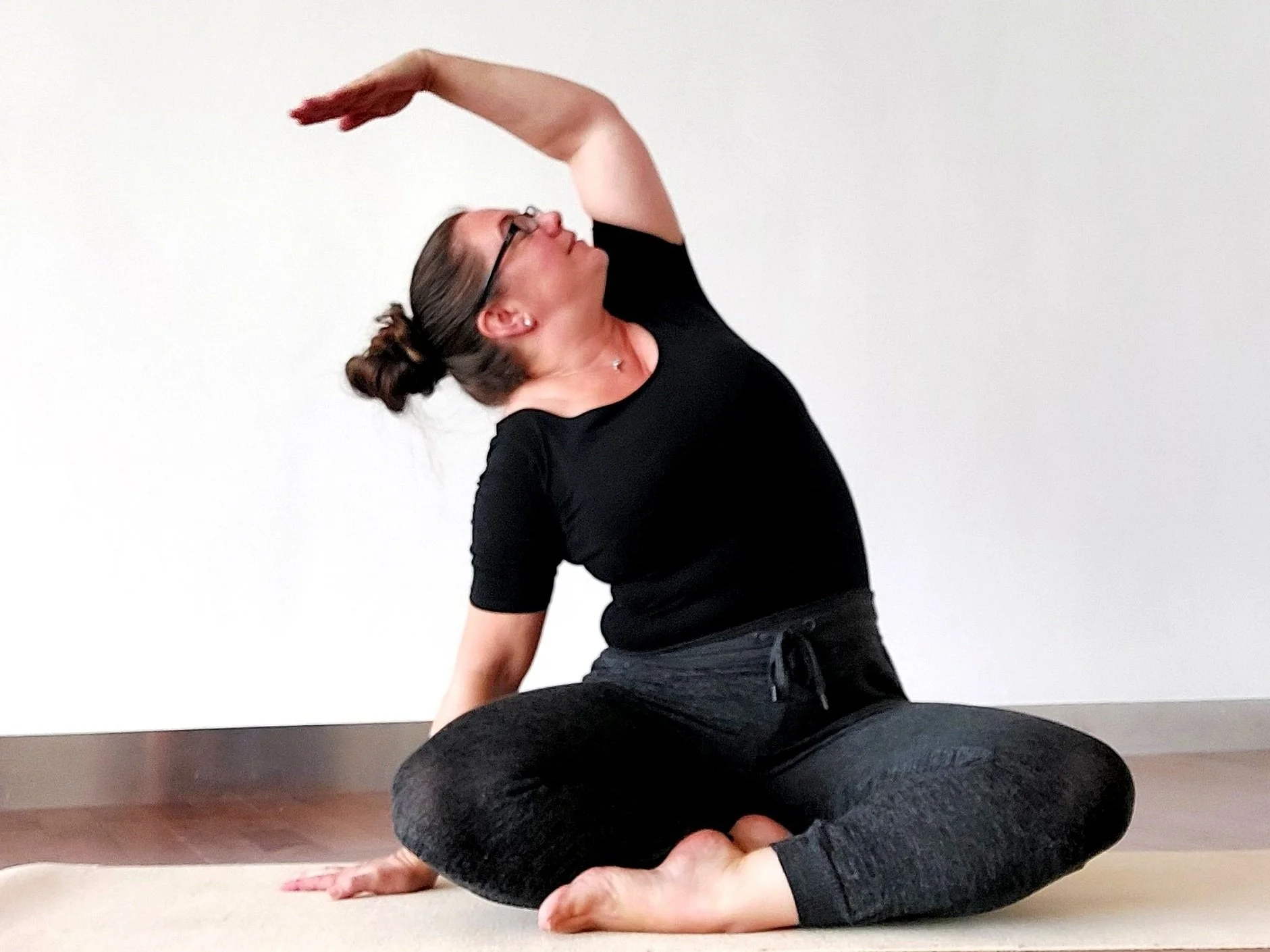 Woman wearing glasses, black bodysuit and leggings sitting on a yoga mat with crossed legs, bending to the side and looking up at left arm raised above the head