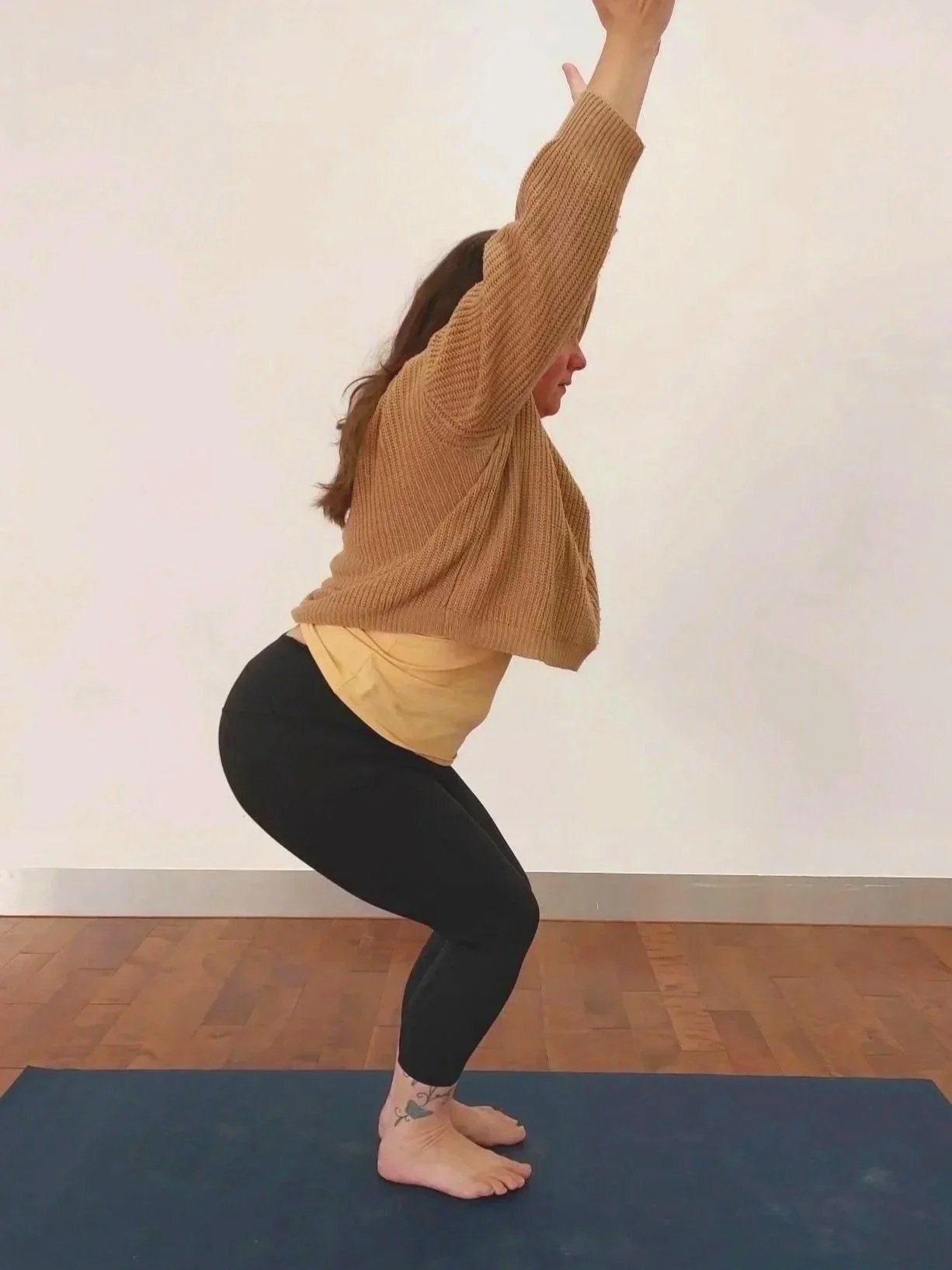 Woman in a beige jumper on a yoga mat in chair pose with arms above the head
