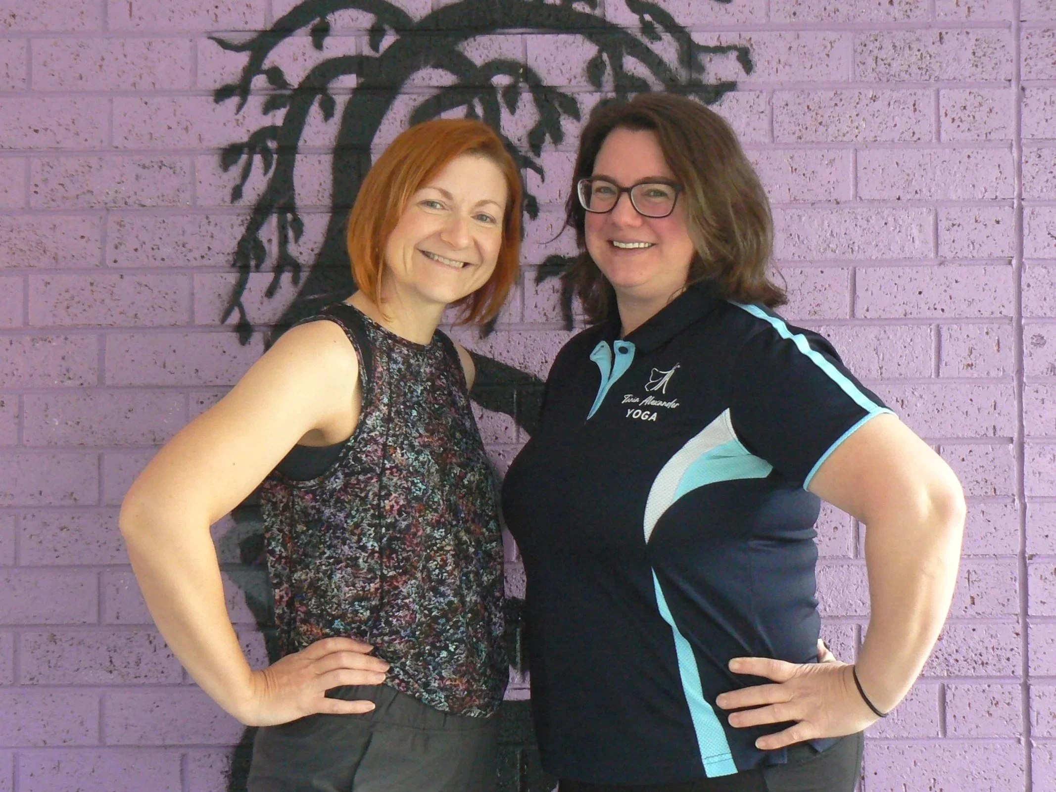 Two white women standing side on to one another with outer hand on hip and turning to face the camera with toothy smiles