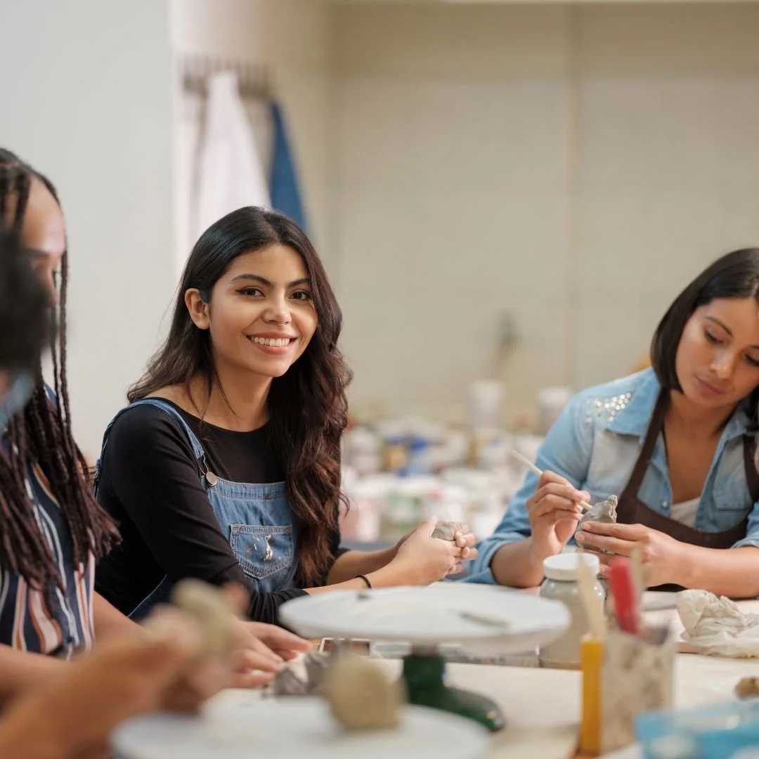 Young woman with long dark hair smiling at the camera while seated at a table full of art supplies with other women