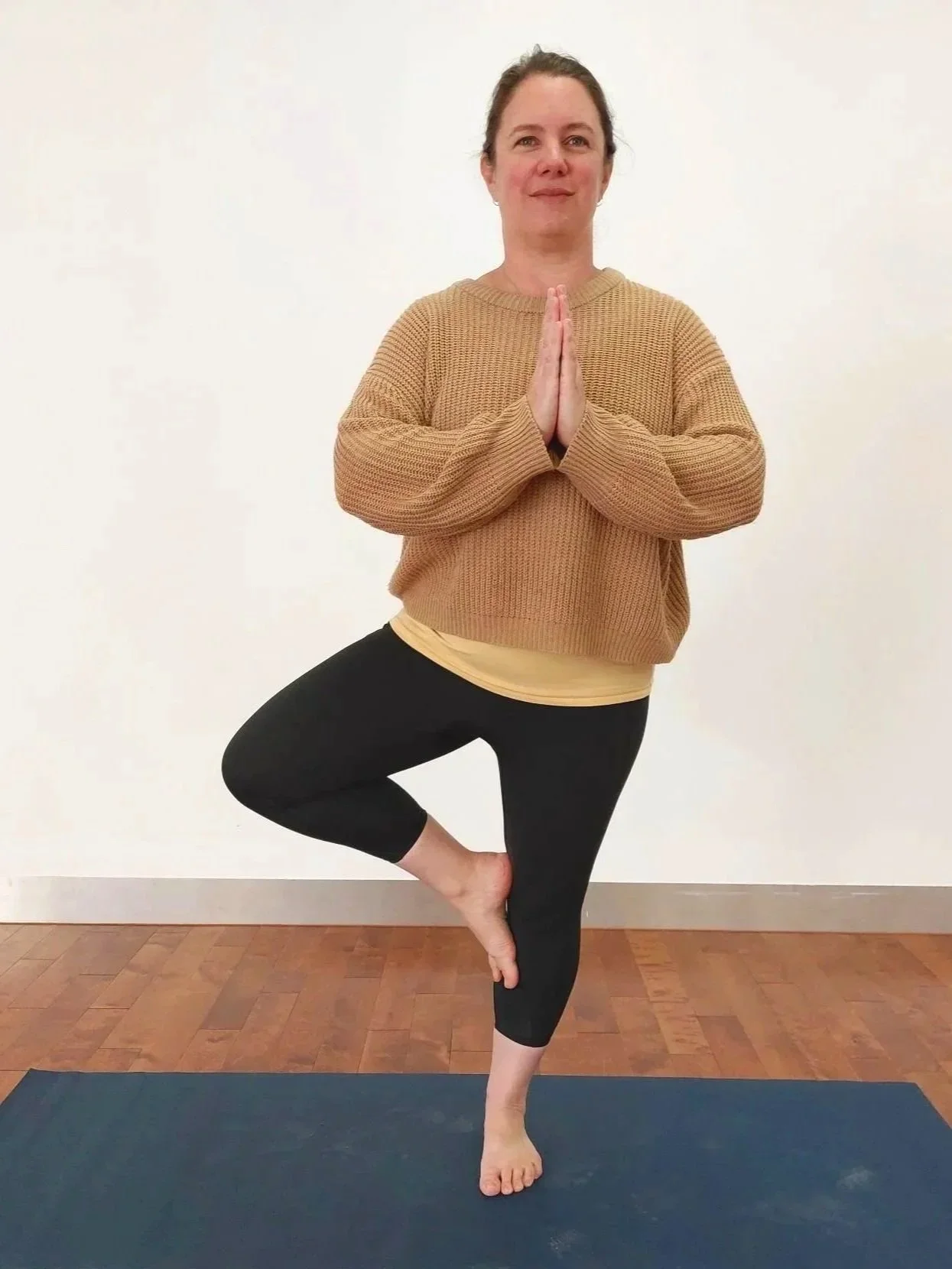 Woman in a beige jumper standing on a yoga mat facing forward in Tree Pose with right knee out to the side and hands in prayer position in front of the chest
