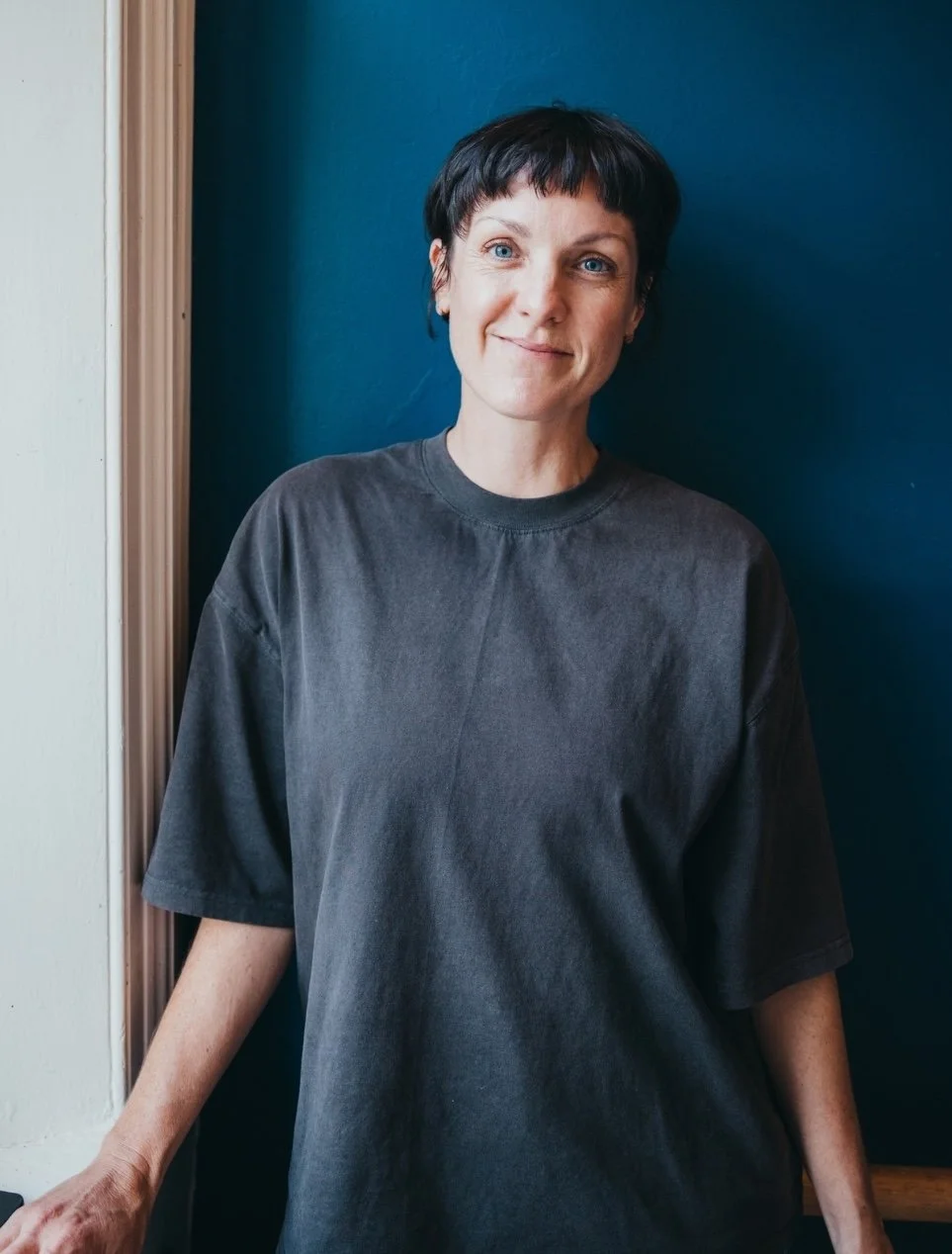 A woman with short dark hair wearing a loose gray T-shirt standing indoors against a dark blue wall with a wooden door frame on her left.
