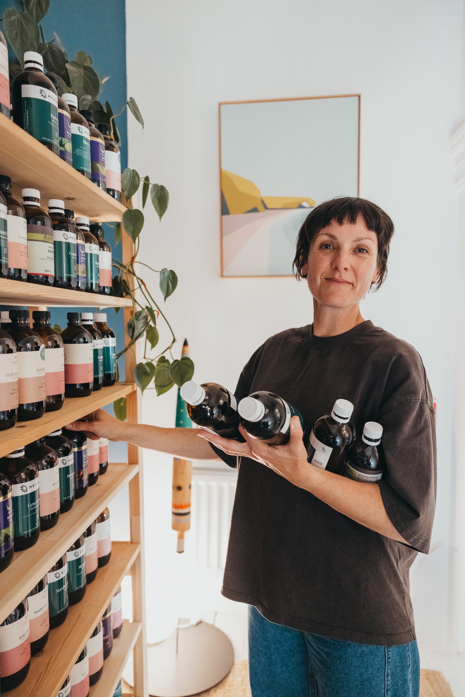 A woman in a dark gray shirt and blue jeans standing near wooden shelves filled with bottles of supplements or vitamins, holding several bottles in her hands, and smiling.