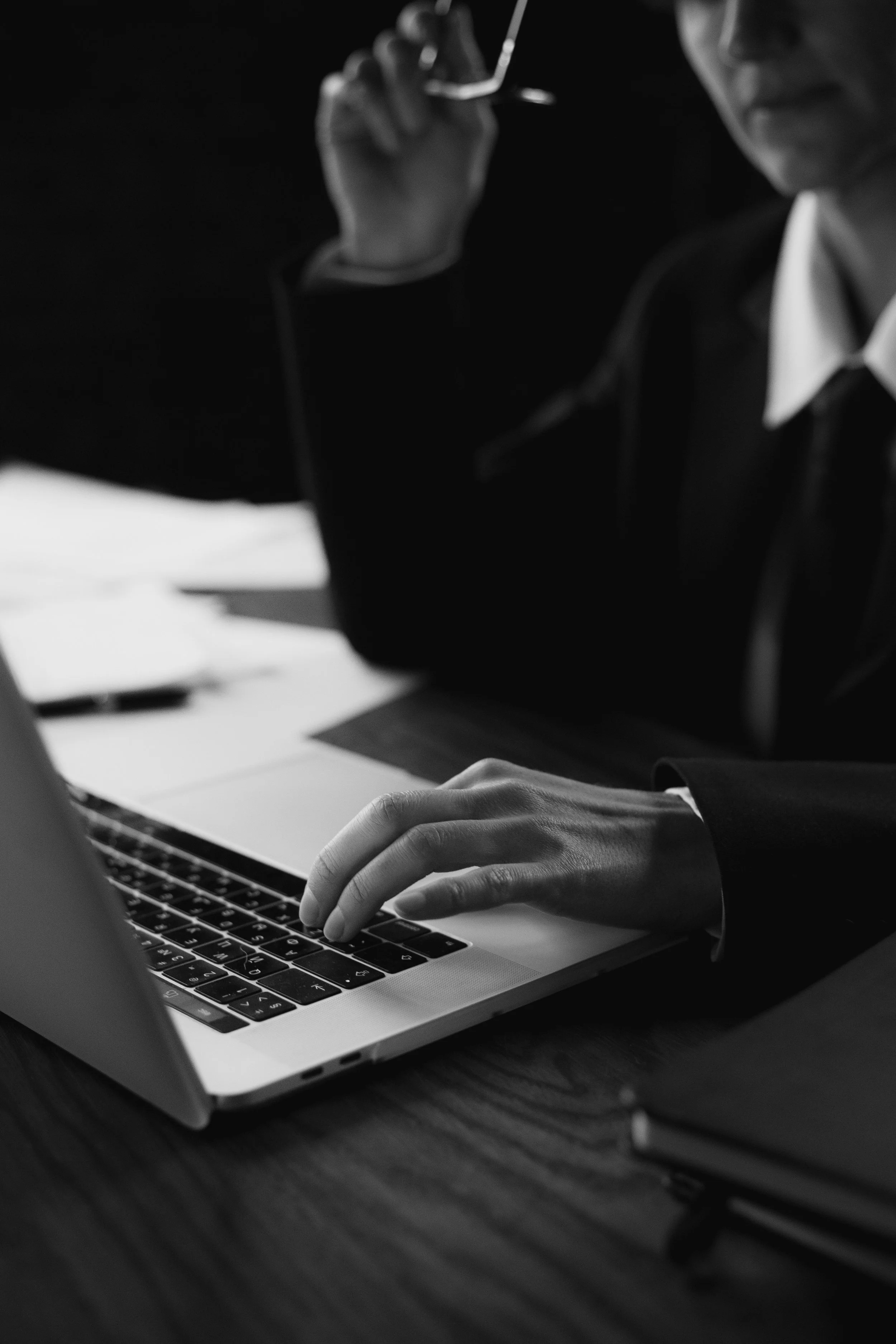 A person in business attire working on a laptop at a desk with documents and notebooks, black and white photo.
