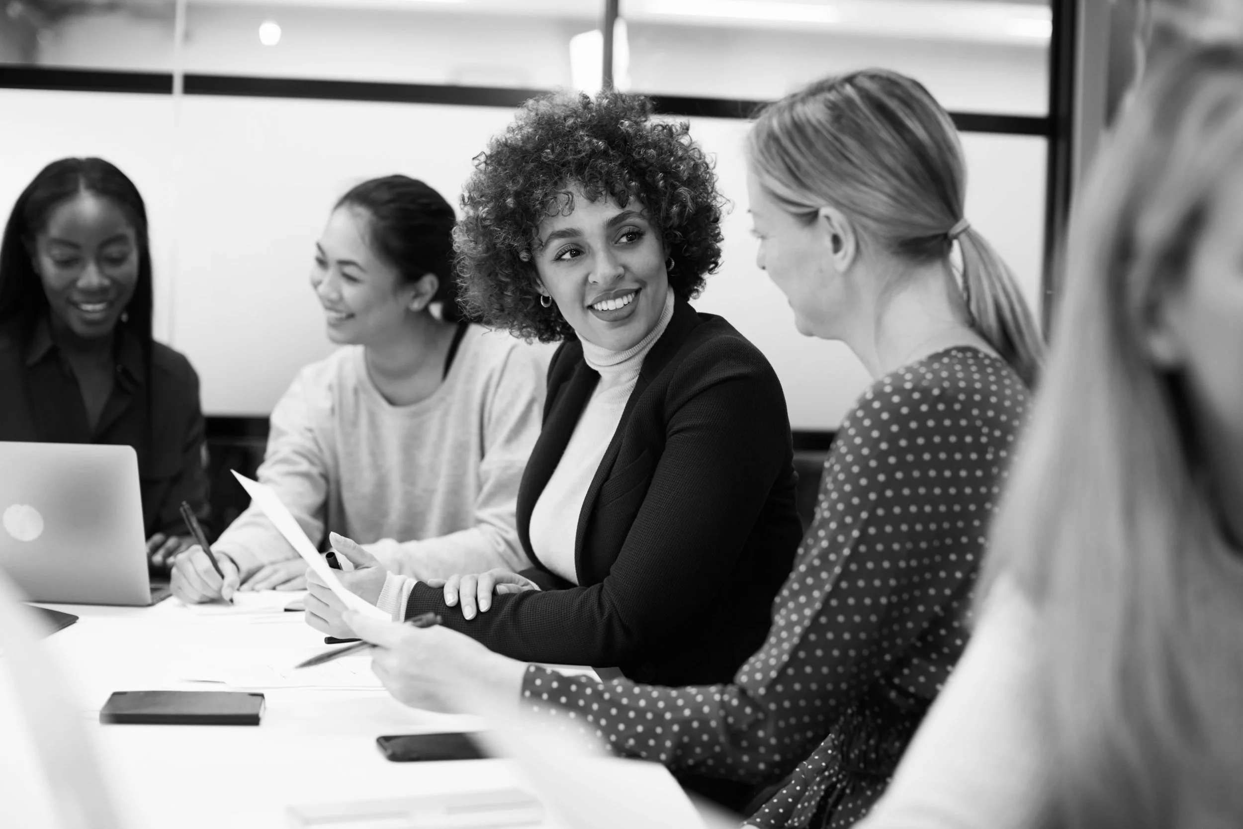 A diverse group of women sitting around a table in a meeting, smiling and engaging in conversation.