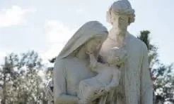 A white marble statue of a woman holding a child, outdoors with trees and a cloudy sky in the background.
