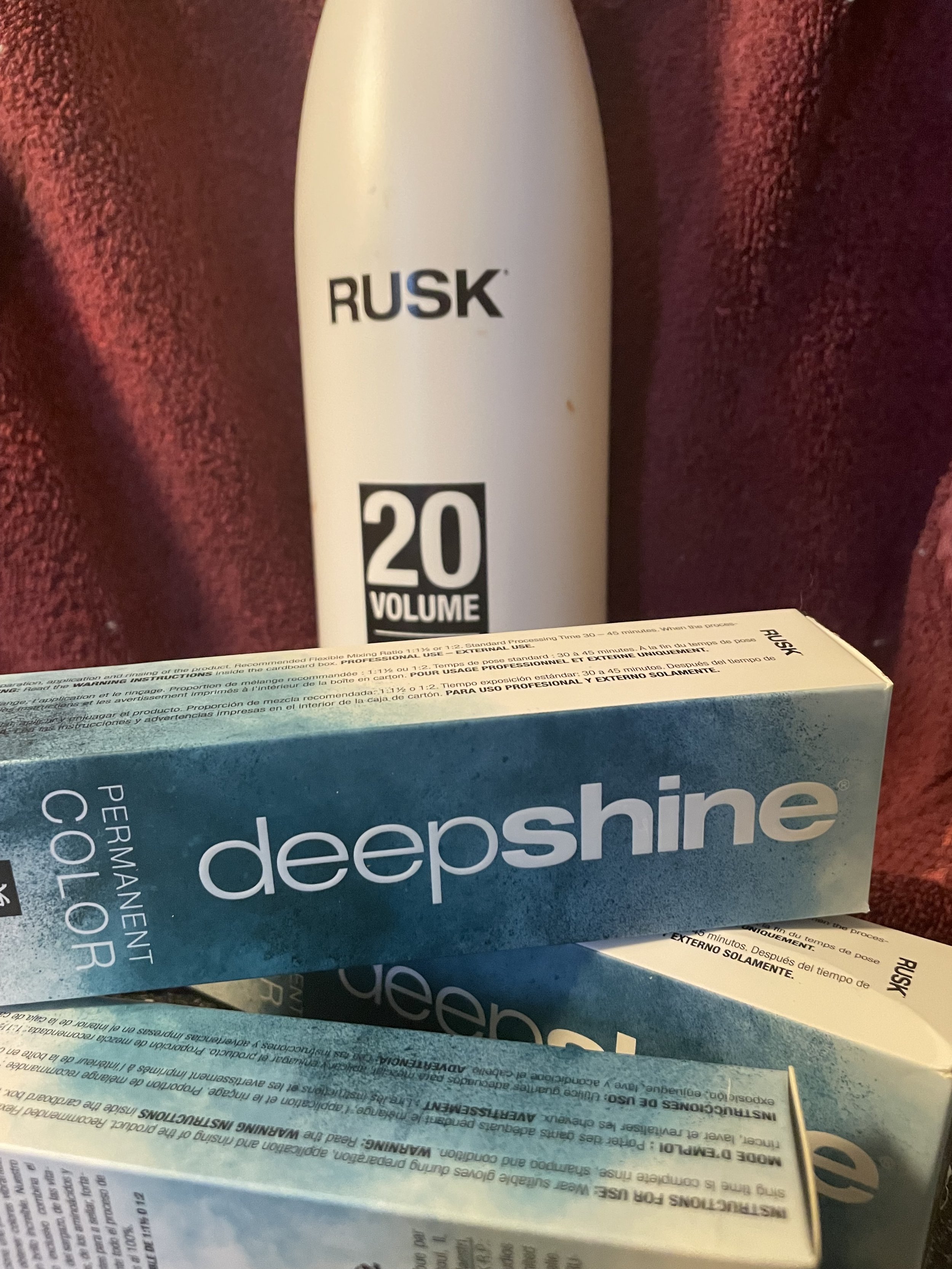 A white bottle of Rusk volume text on it, positioned above a blue box labeled Deepshine professional color spray, with warning instructions on the box. The background is a red textured fabric.
