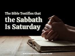 A Bible resting on a wooden surface with a hand in a praying position beside it, against a black background with white text that reads, 'The Bible Testifies that the Sabbath Is Saturday.'