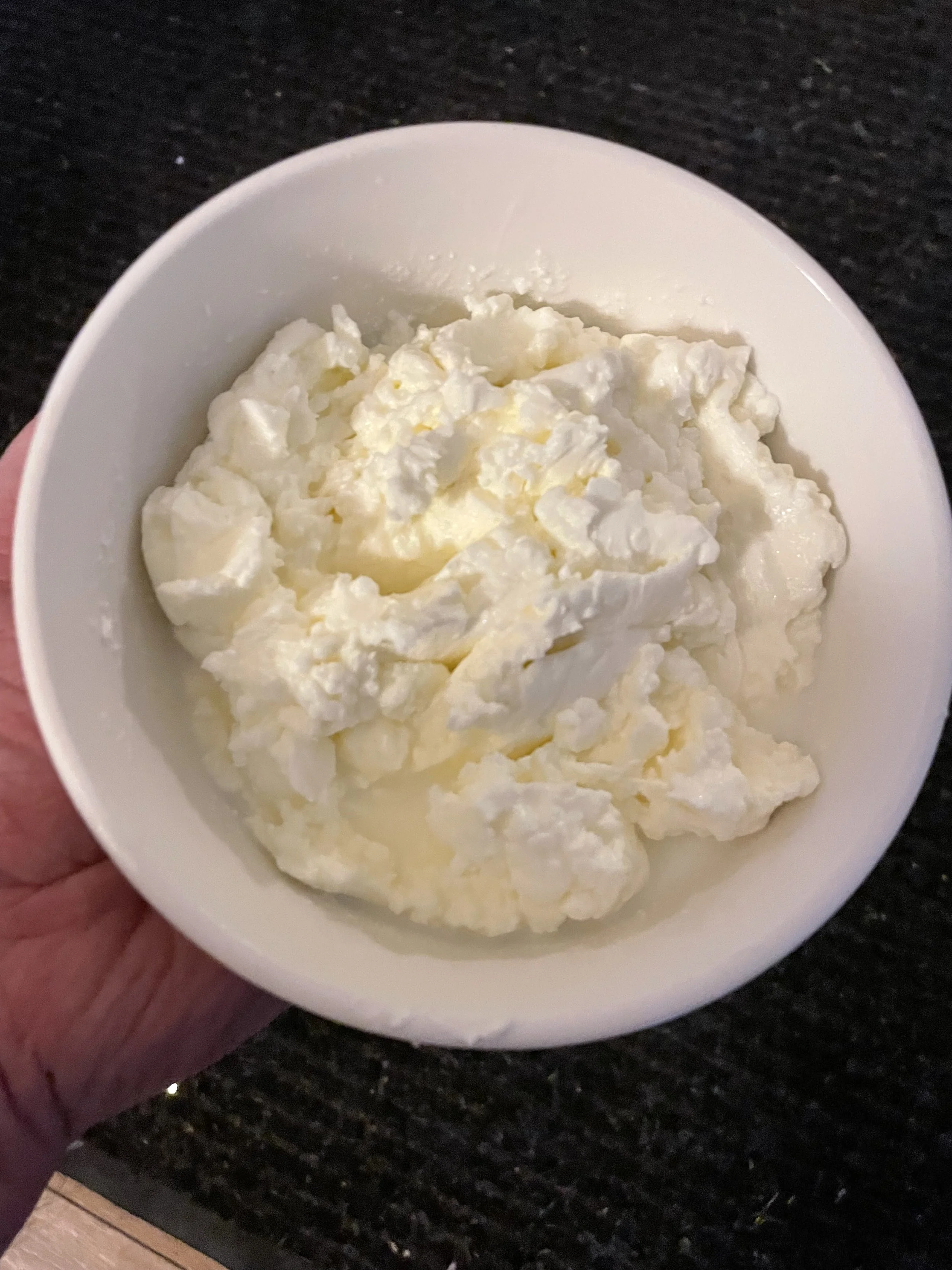 A white bowl filled with homemade butter, being held by a hand with a dark granite countertop in the background.