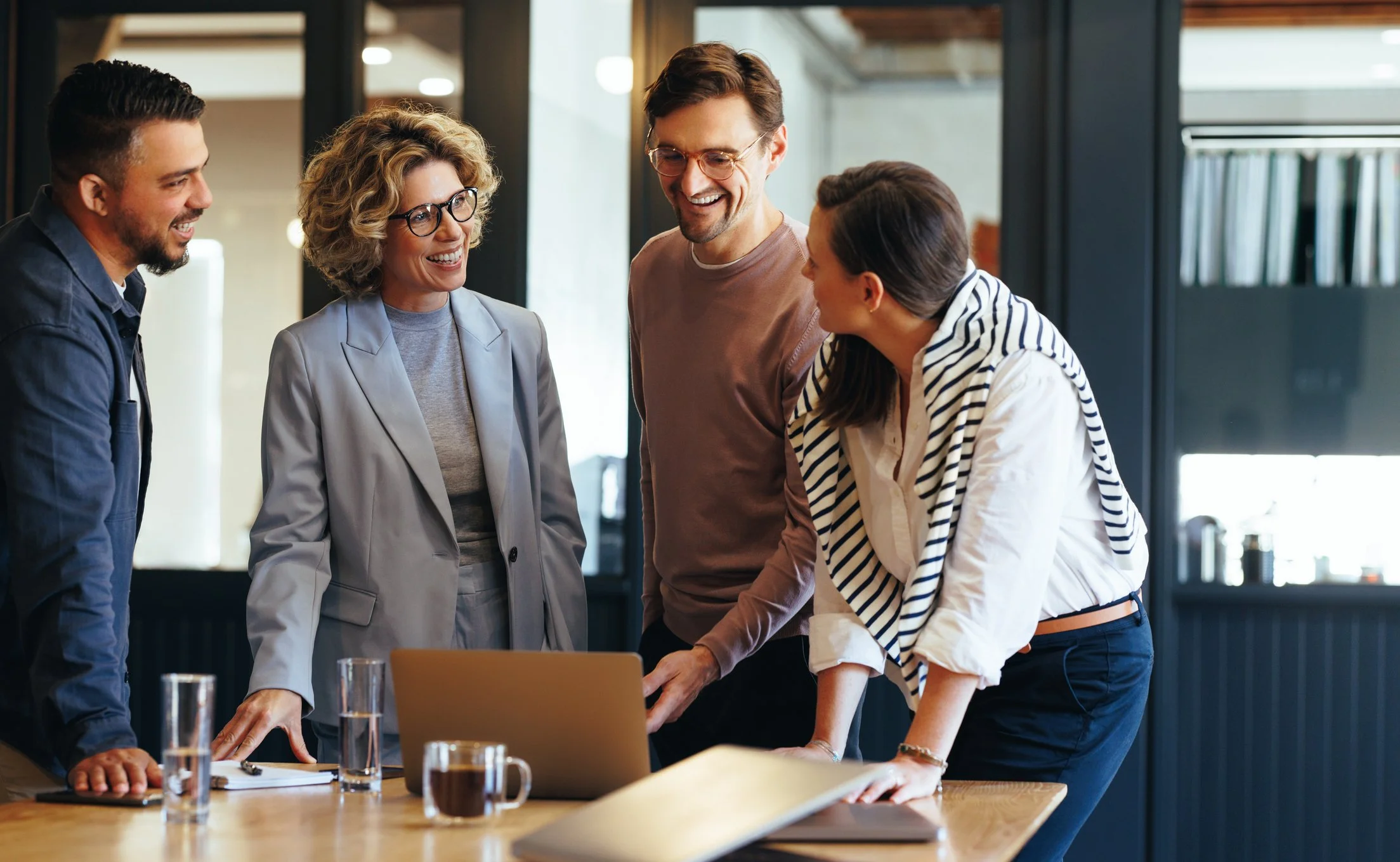 A group of five diverse professionals gathered around a table in an office, smiling and engaged in a conversation, with a laptop and documents on the table.