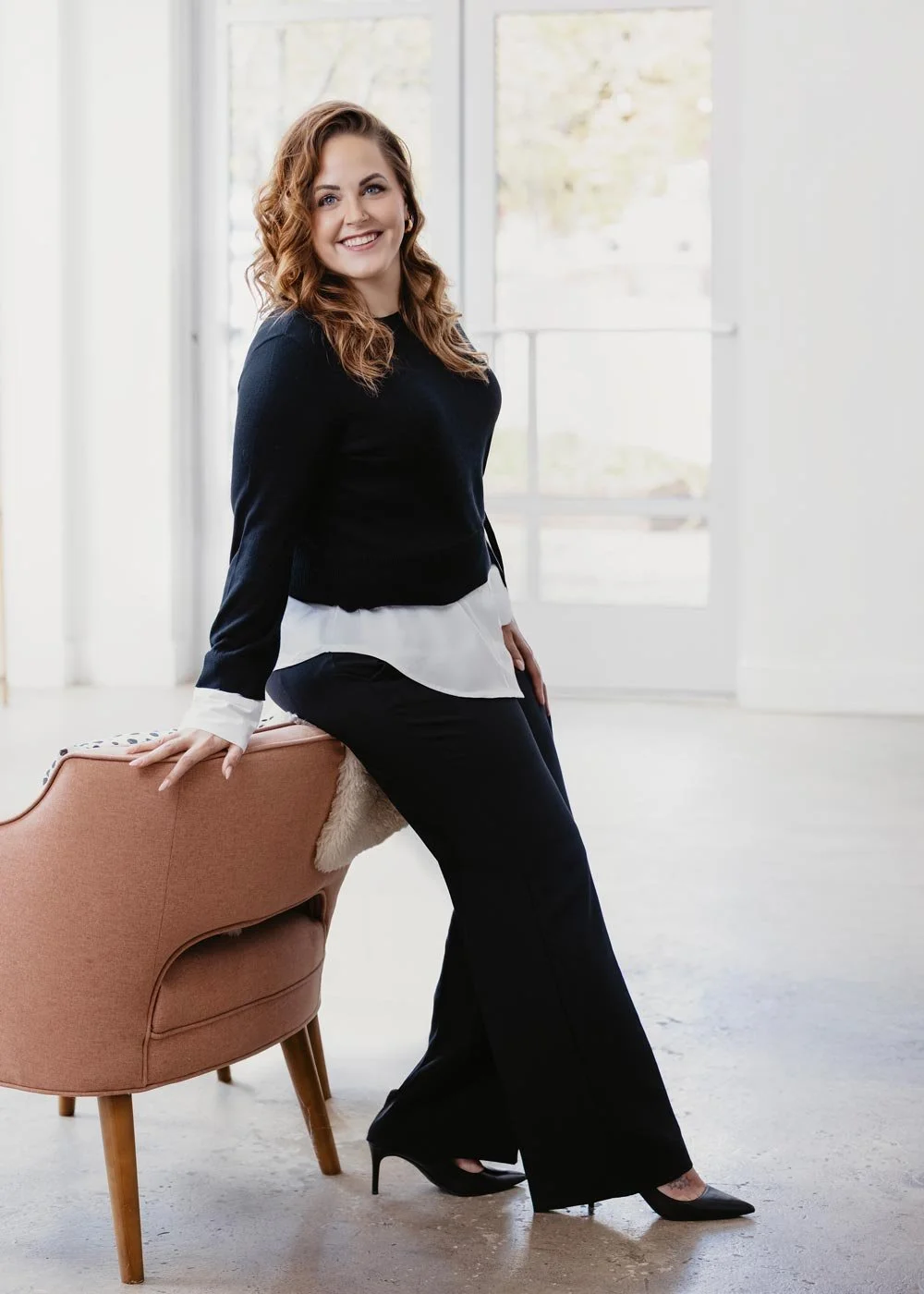 A woman with curly brown hair and blue eyes sitting on a pink armchair, smiling, in a bright room with large windows, dressed in a black sweater, white shirt, black pants, and black heels.