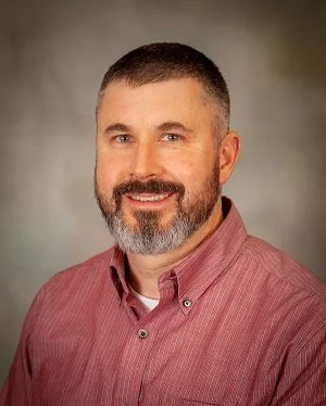 Portrait of a bearded man with short dark hair, wearing a red button-up shirt, smiling against a neutral background.