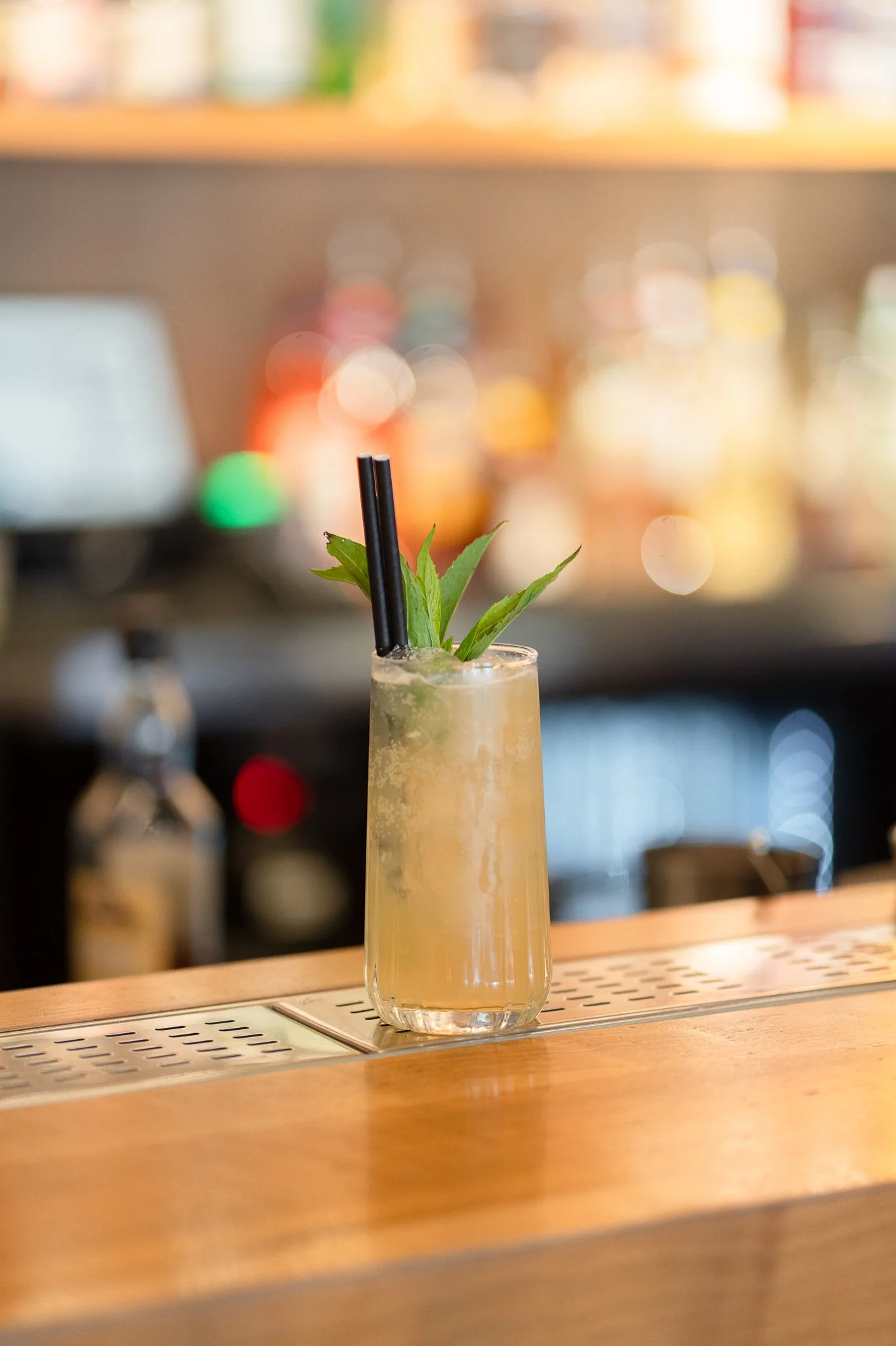 A tall, clear glass cocktail with a yellowish beverage, garnished with green leaves and two black straws, placed on a wooden bar counter.