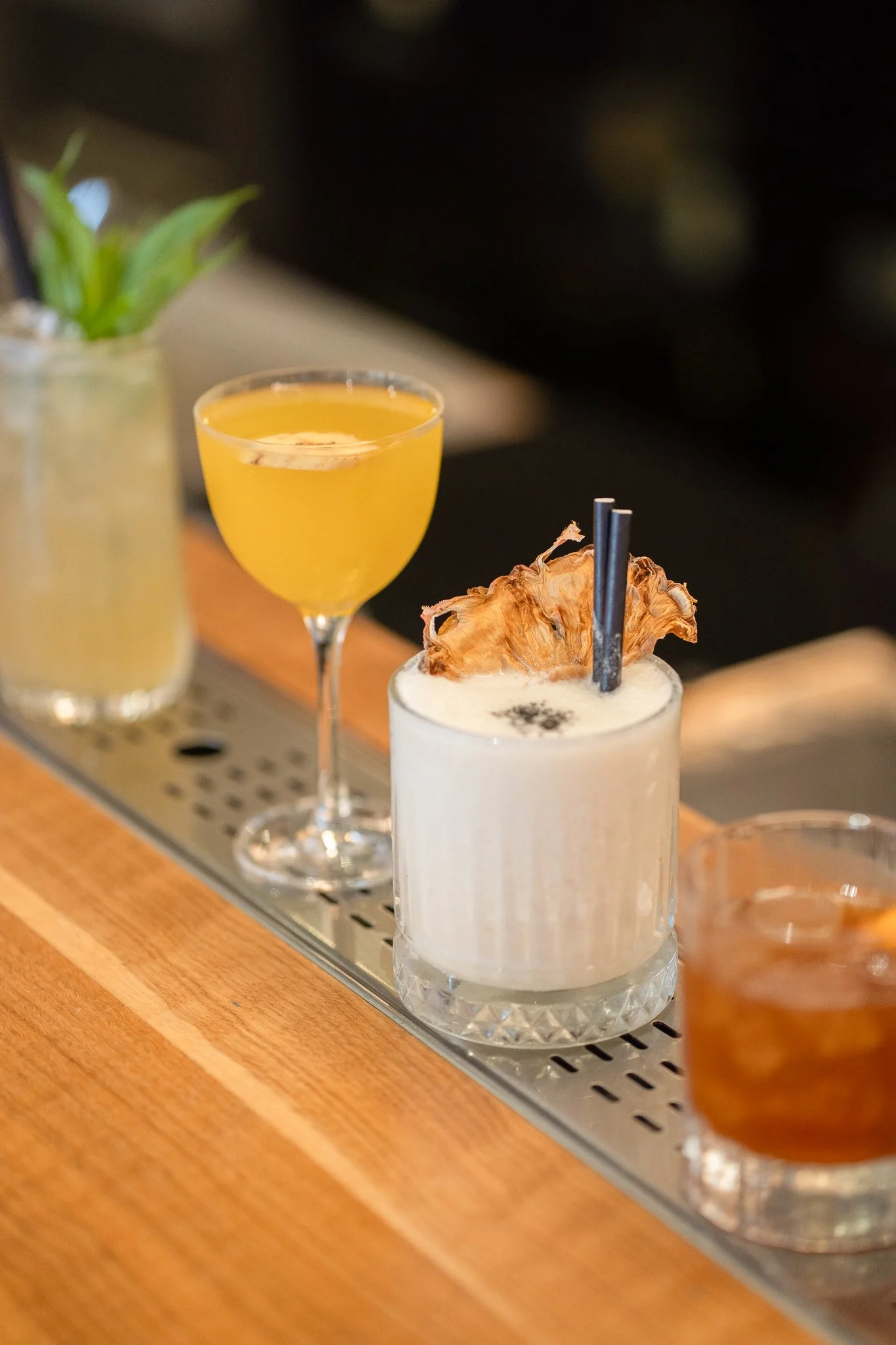 A row of various cocktails on a wooden bar with a plant in the background.
