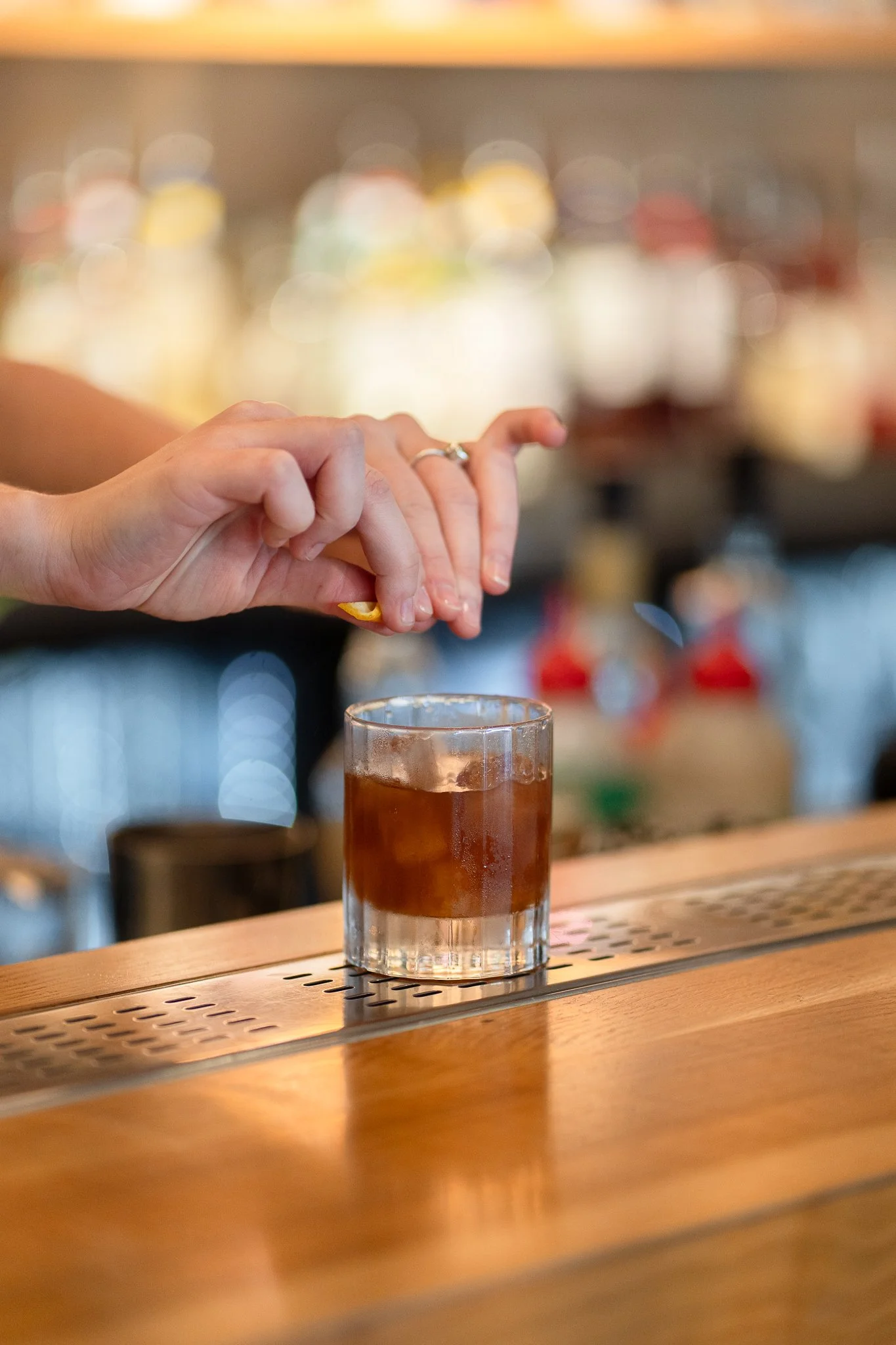 A person's hand placing a lemon twist garnish on a cocktail in a shot glass on a bar counter.
