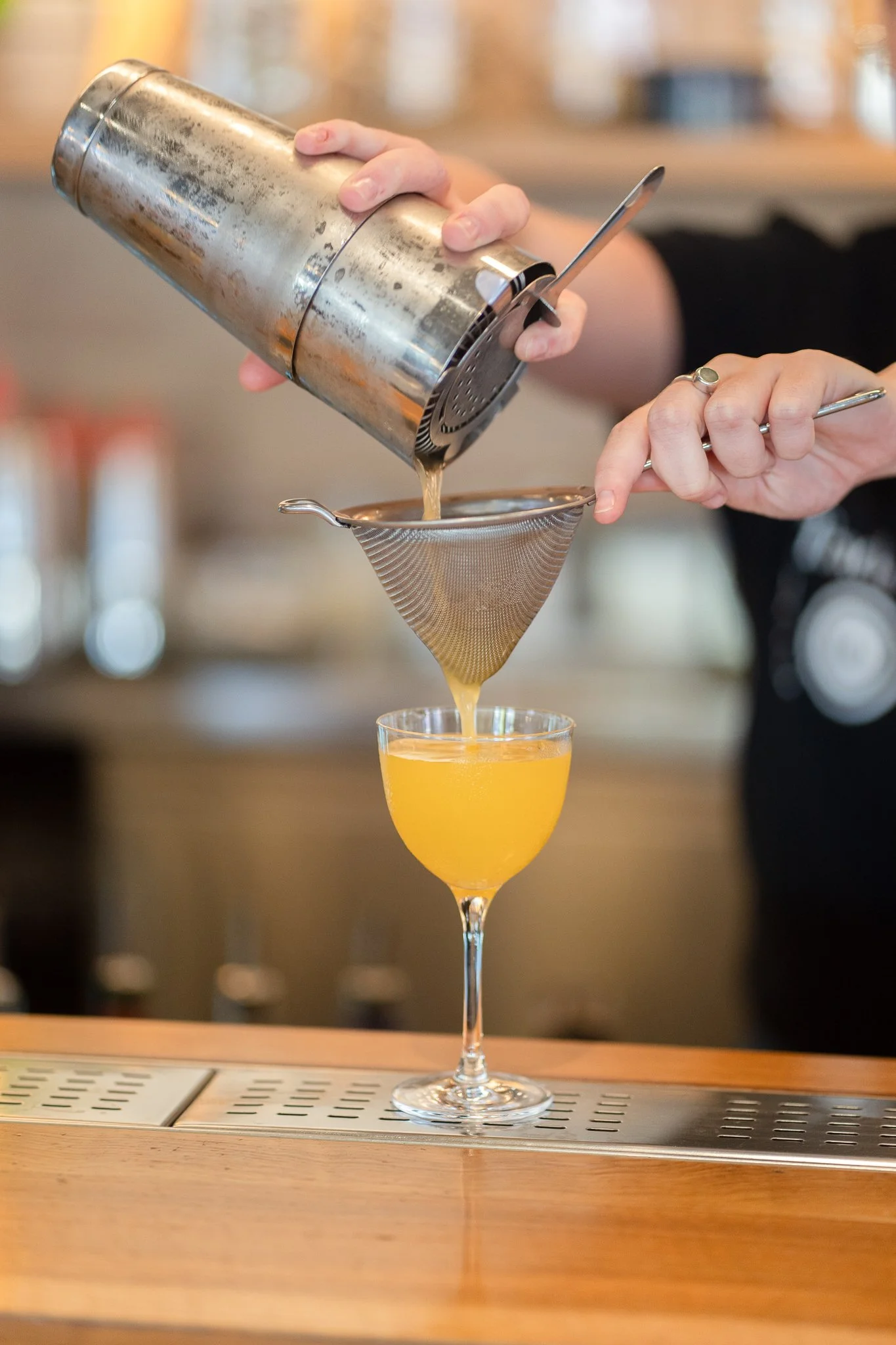 A bartender pours a yellow-orange drink through a metal strainer into a glass with a stem. The bartender is using a shaker and a spoon, and the background is a bar setting.