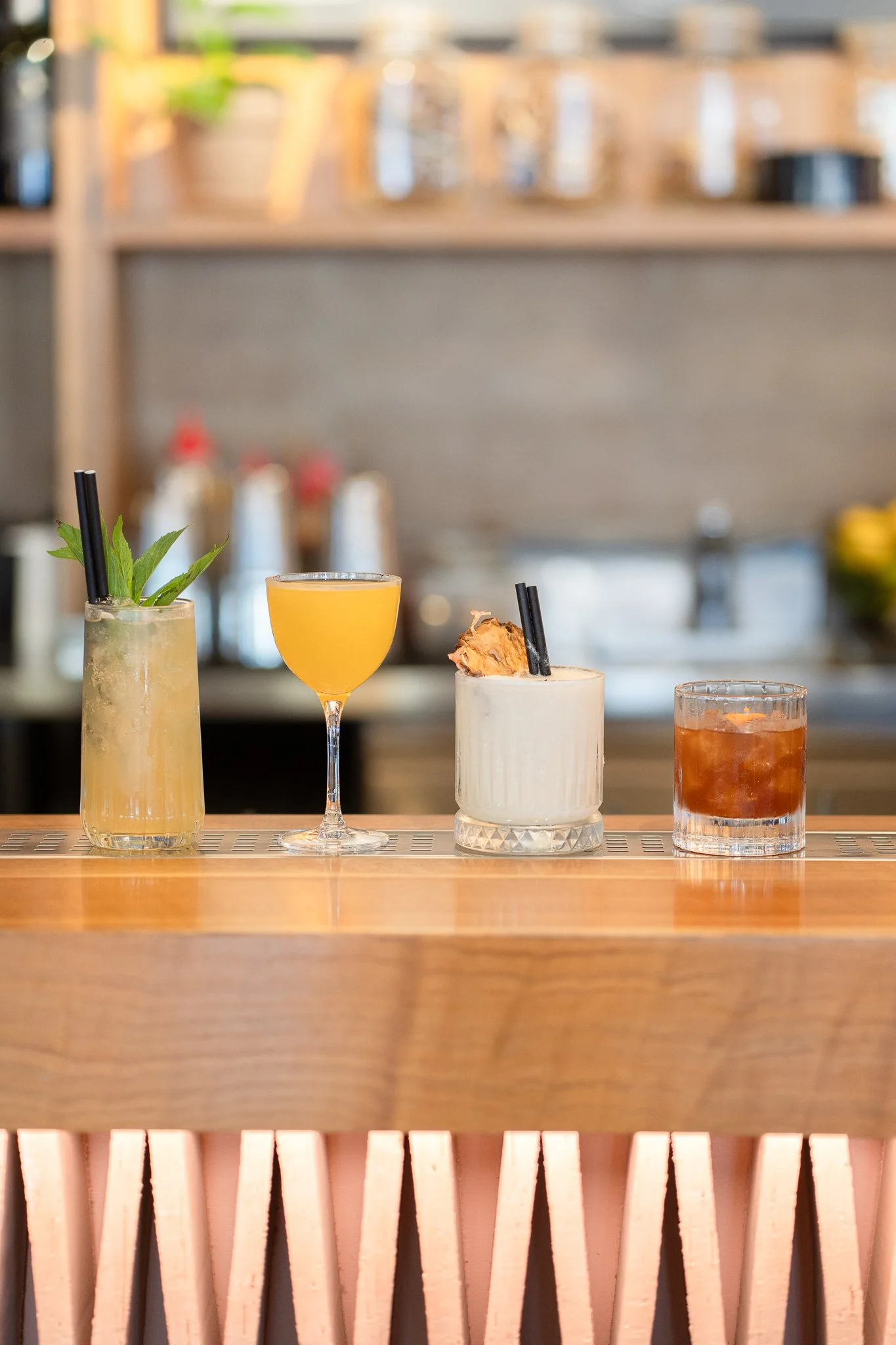 Four colorful cocktails on a bar counter with a wooden front panel and a blurred background of shelves and bottles.
