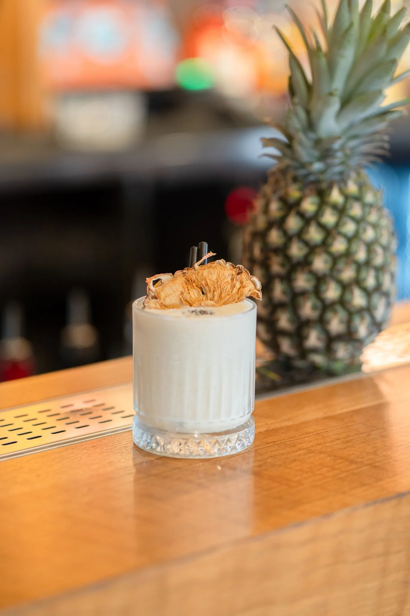Close-up of a cocktail in a transparent glass with a pineapple slice garnish, placed on a wooden bar counter with a pineapple in the background.