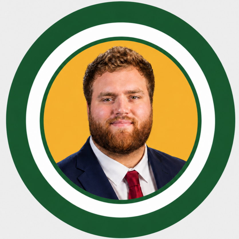 A young man with a full beard and curly hair, wearing a suit and tie, smiling, with a yellow background.